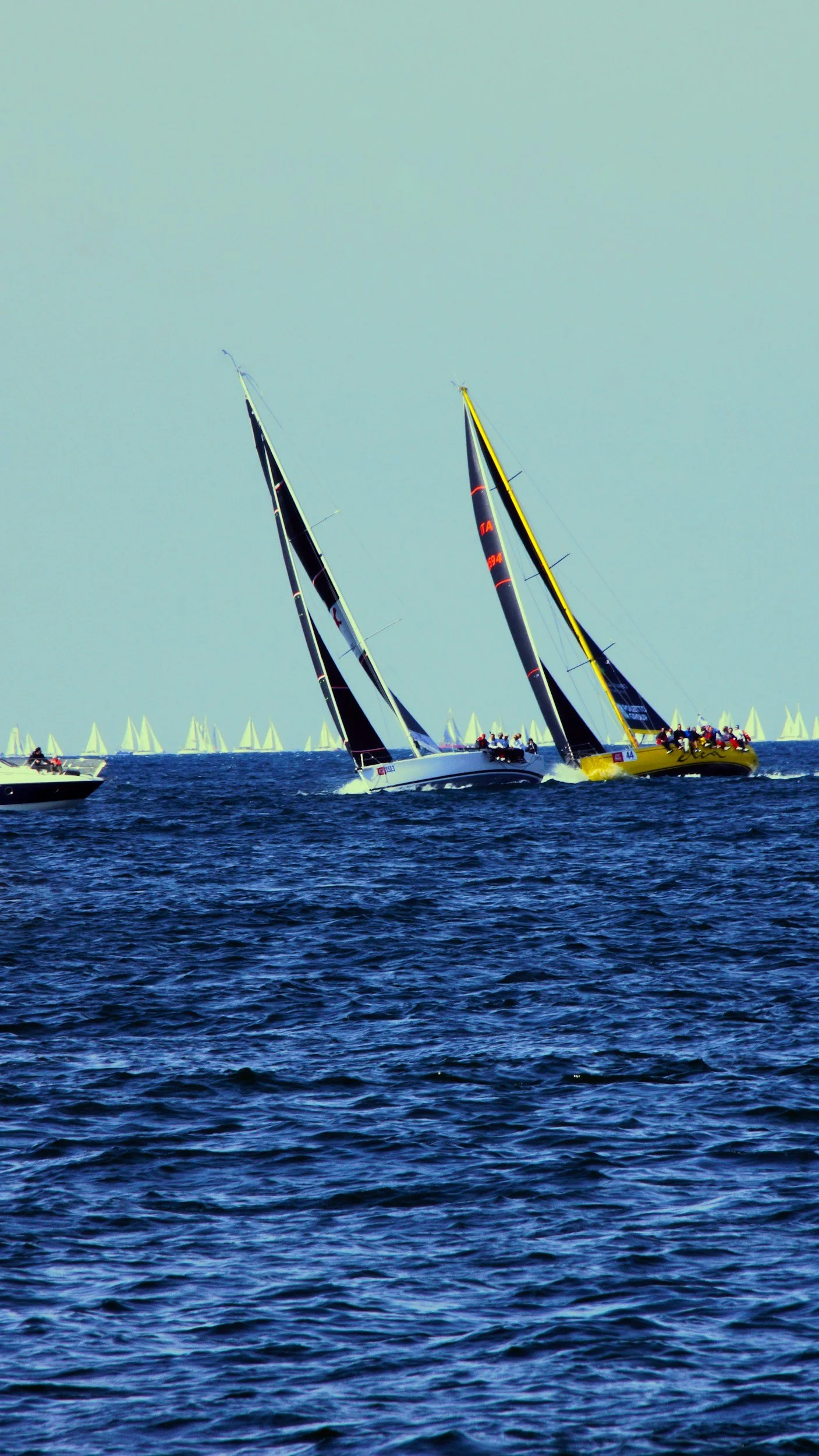 Two sailboats racing on the ocean with other sailboats visible in the background.