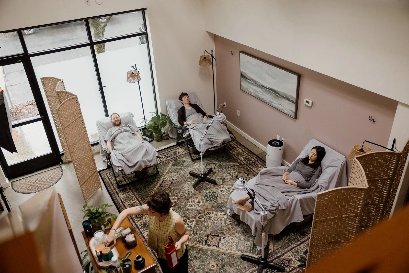 Overhead image of a calm healing space where three patients rest quietly in zero-gravity chairs while covered in blankets. Acupuncturist Kelly Baucum prepares for the next treatment in the back of the room.