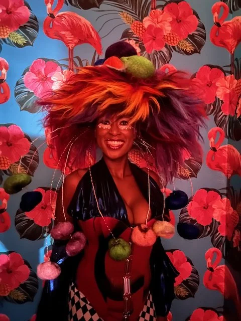 Woman with a big colorful feathered hat and vibrant outfit standing in front of a tropical flamingo and hibiscus flower patterned background.