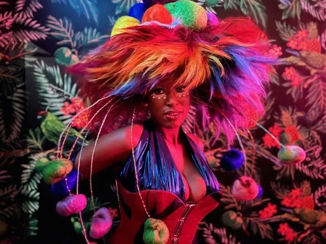 Person wearing a vibrant, multicolored headdress with fuzzy, fluffy textures, and a revealing metallic outfit, standing against a tropical-themed background.