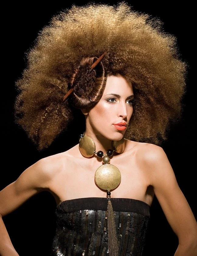 A woman with large, voluminous curly hair styled with an hair stick and bun on top and makeup with red lipstick, wearing a strapless black sequined dress and a large gold necklace with beads and chains, against a black background.