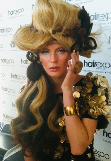 Woman with elaborate, voluminous blonde hair styled with large black and brown curlers, wearing black and gold clothing with floral embellishments, gold cuff bracelet, posing in front of a hair expo backdrop.