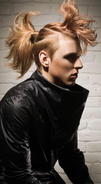 Side profile of a young woman with dramatic, spiked hairstyle in light brown and blonde, wearing a black leather jacket, posed against a white brick wall.