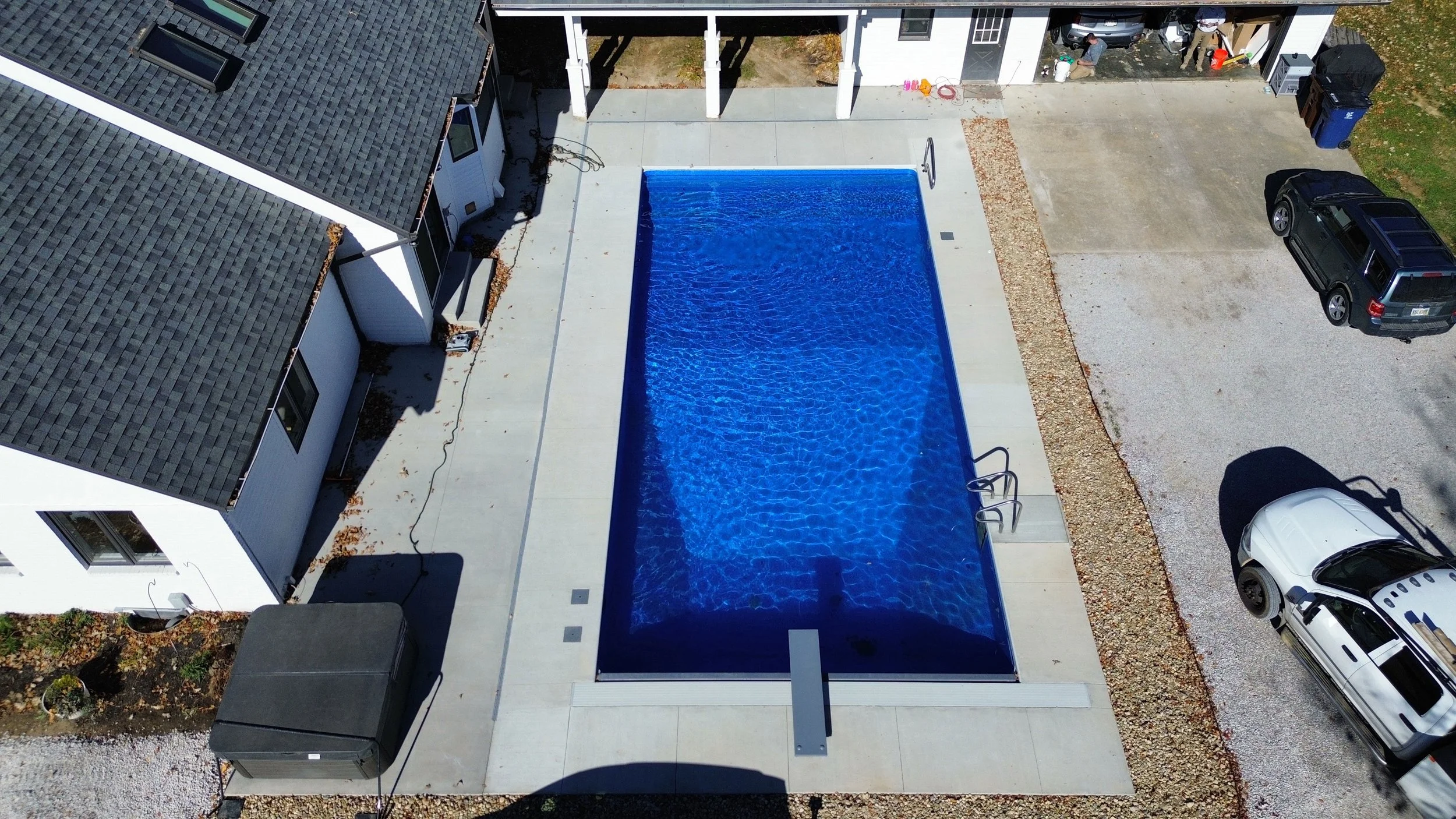 Aerial view of a backyard with a rectangular swimming pool, surrounded by a concrete patio, adjacent garage with open doors, and a gravel driveway with parked cars. The garage has four people near it, and outdoor toys and equipment are also visible.
