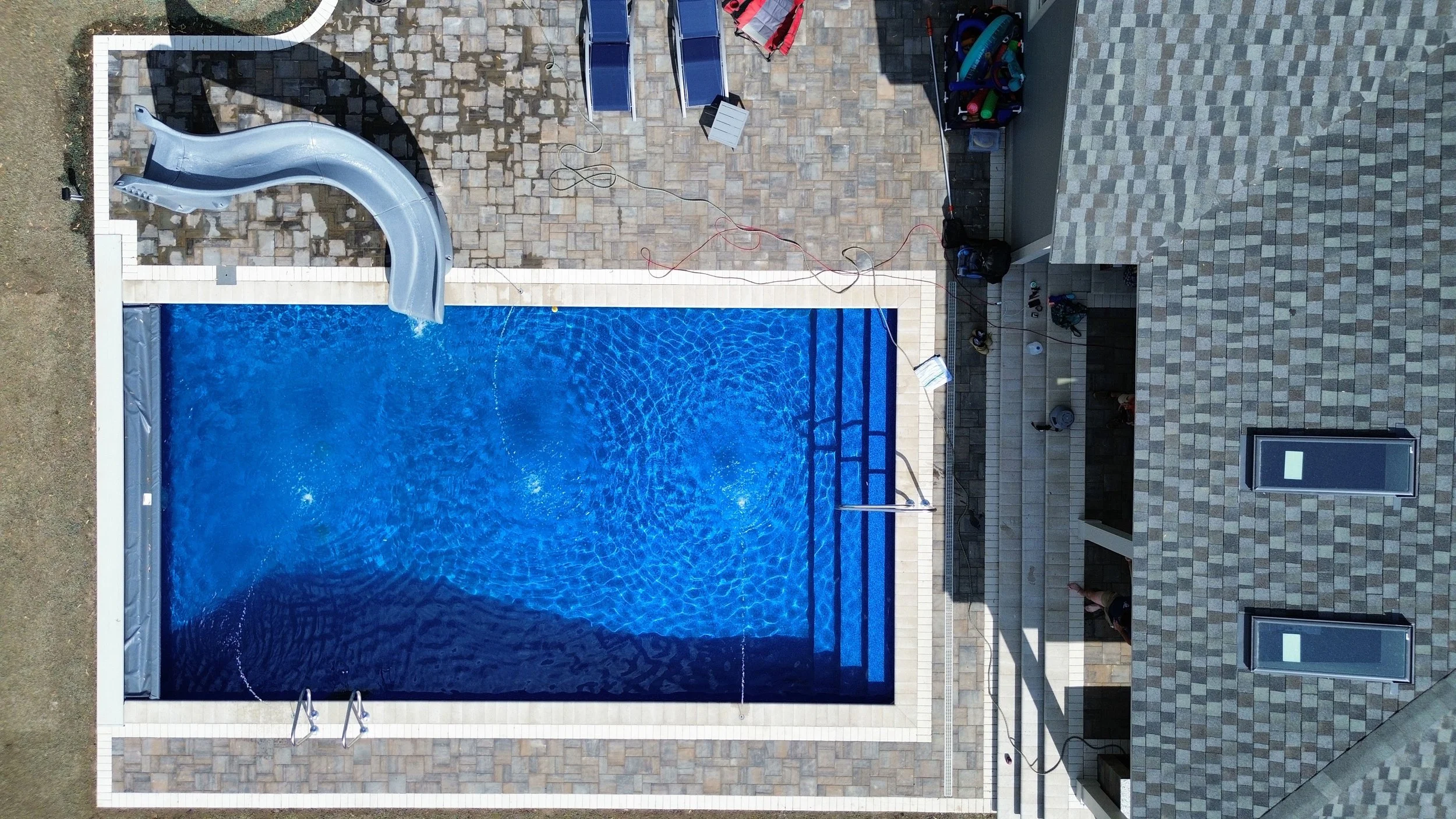 Aerial view of a rectangular swimming pool with a slide, surrounded by a stone-paved patio. There are two blue lounge chairs and some equipment near the pool.