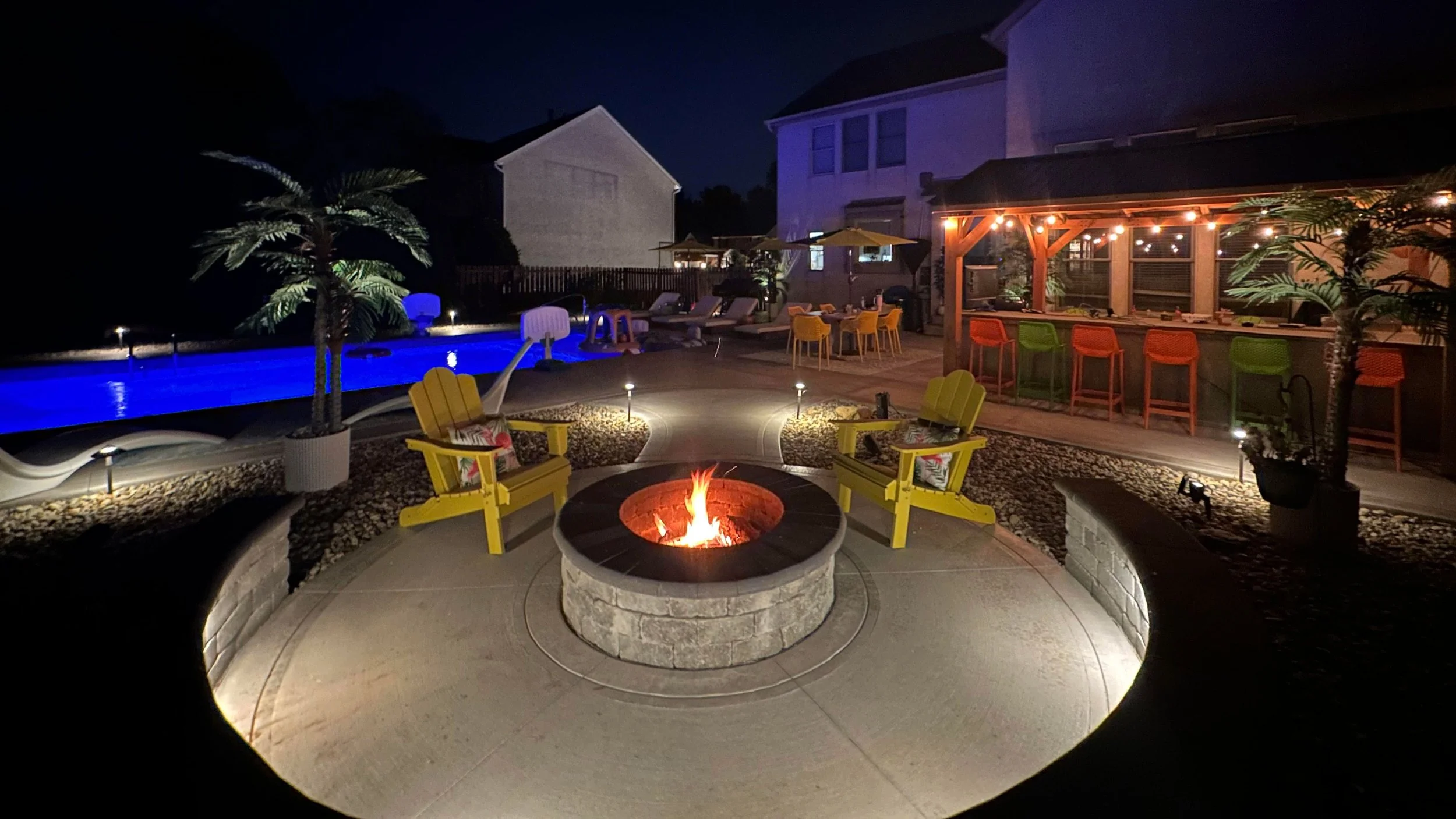 Night view of a backyard with a swimming pool, fire pit, outdoor seating, and a bar, illuminated with colorful lights.