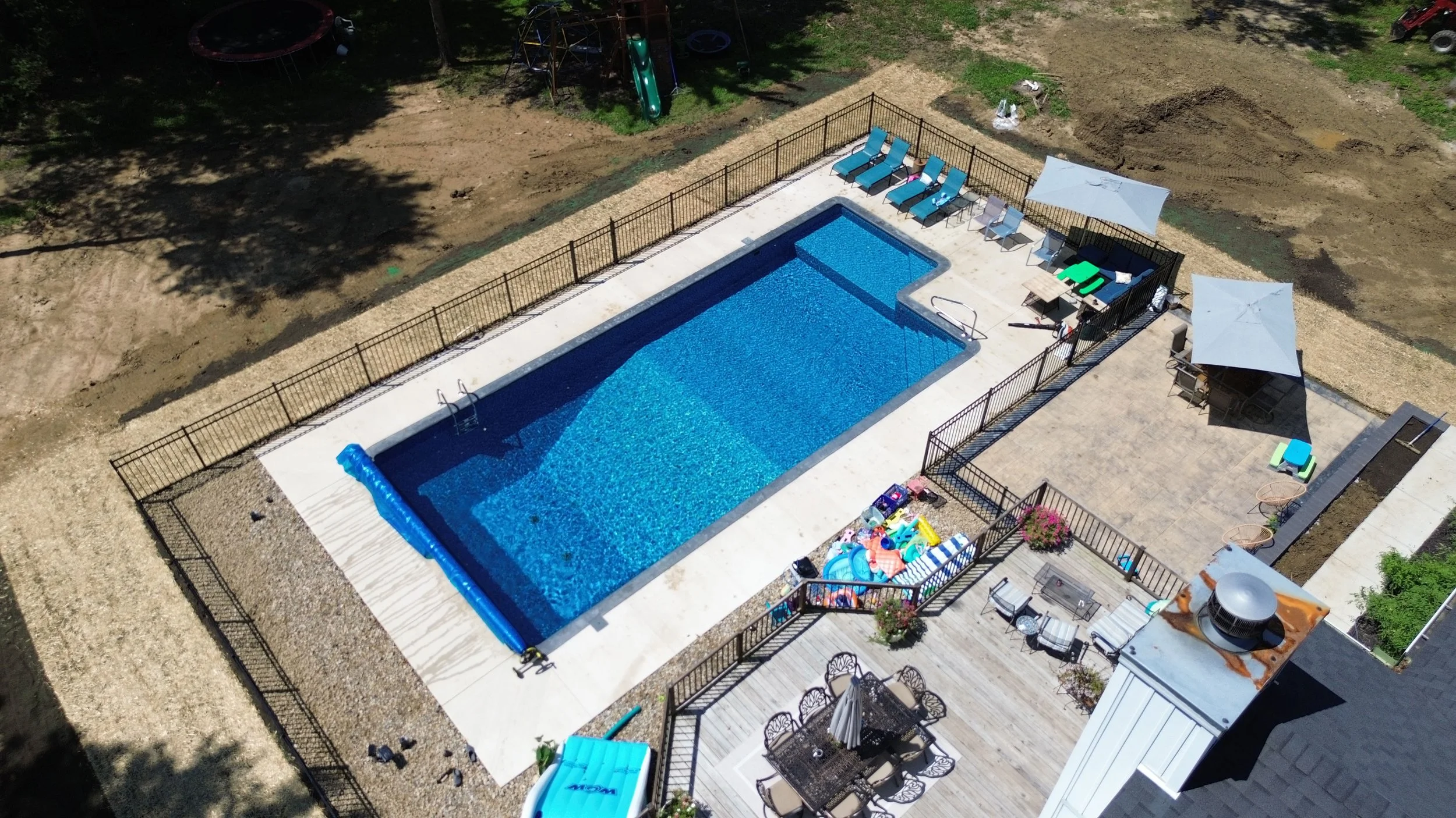 Aerial view of a backyard pool area with lounge chairs, umbrellas, and outdoor furniture, fenced off from surrounding dirt yard and greenery.