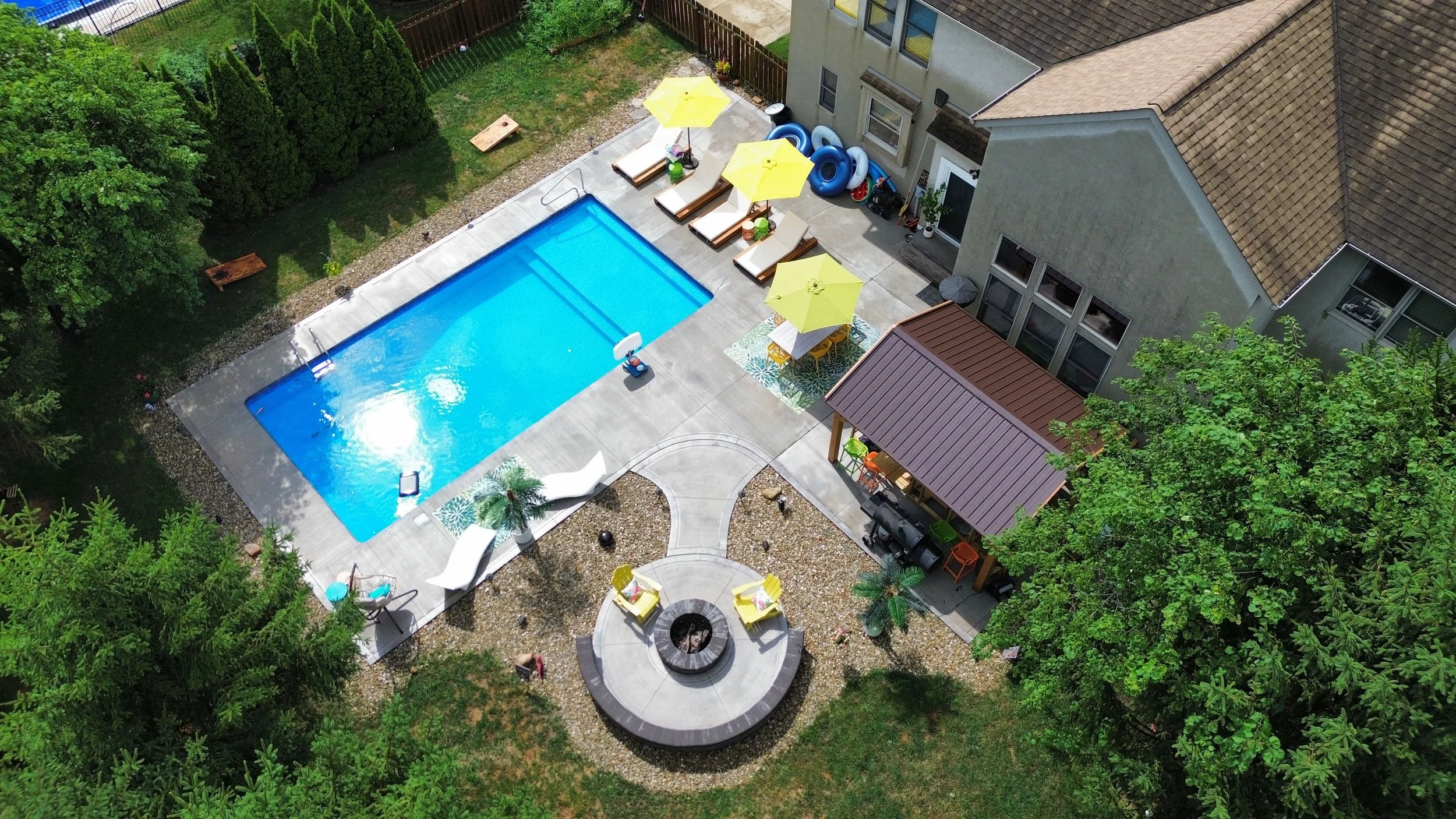 Aerial view of a backyard with a swimming pool, poolside lounge chairs with yellow umbrellas, a covered seating area, a fire pit area with chairs, and lush green trees surrounding the space.
