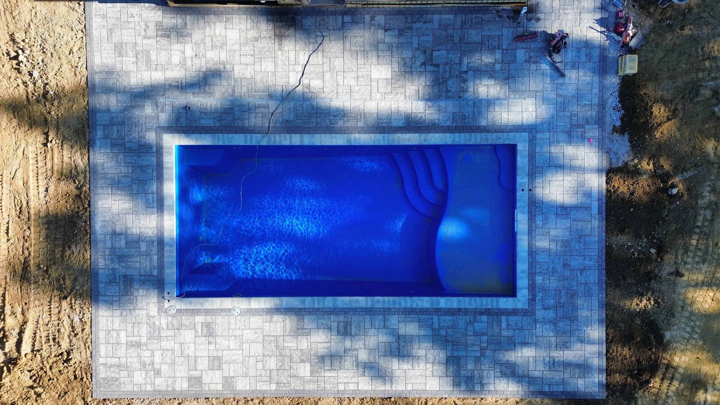 Completed installation of a modern rectangular fiberglass inground pool with a built-in tanning ledge and smooth blue finish, surrounded by a custom stone paver patio.