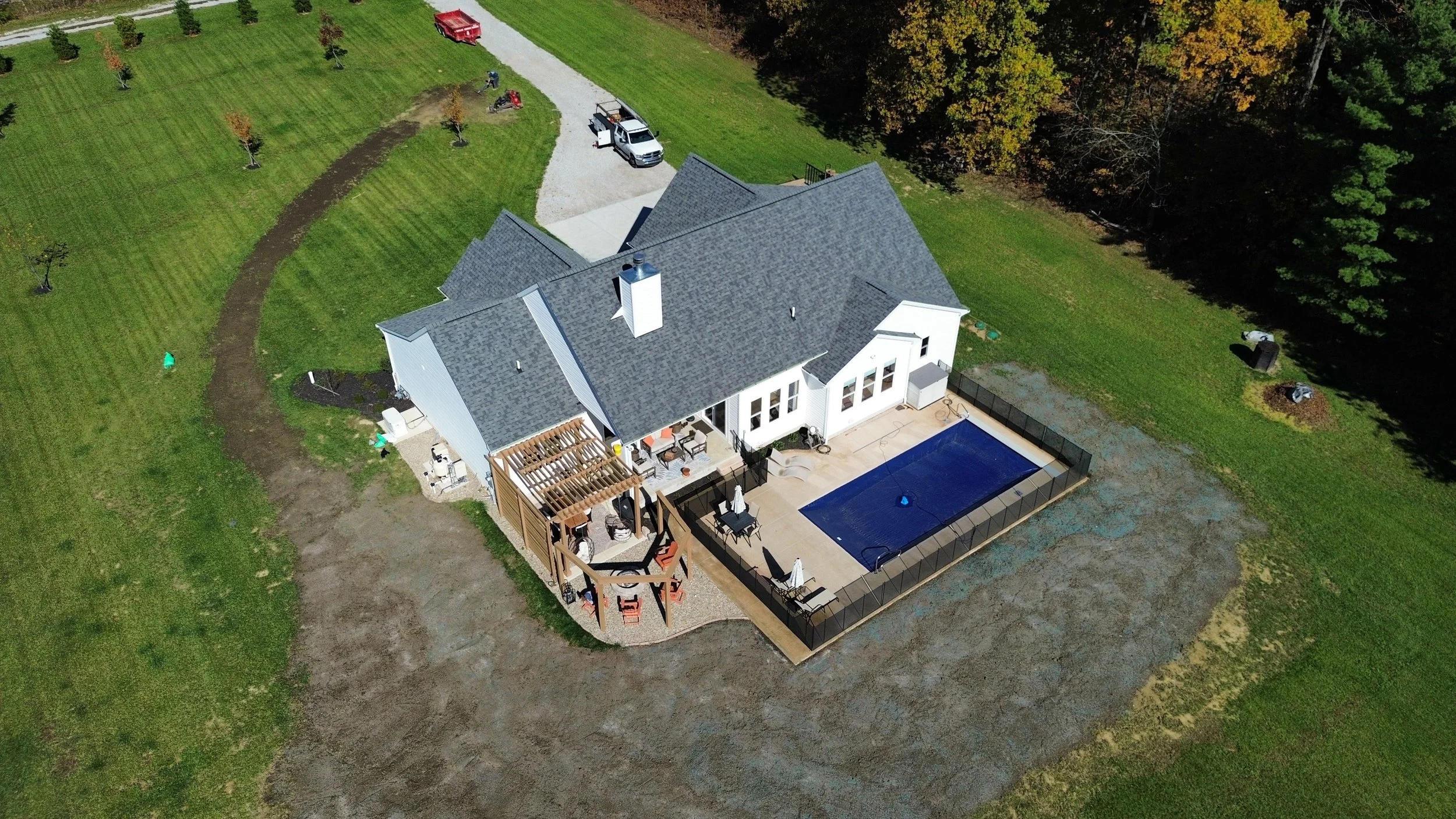 Aerial view of a house with a gray roof, backyard with swimming pool, patio, outdoor furniture, and a wooden deck, surrounded by green lawns and trees.