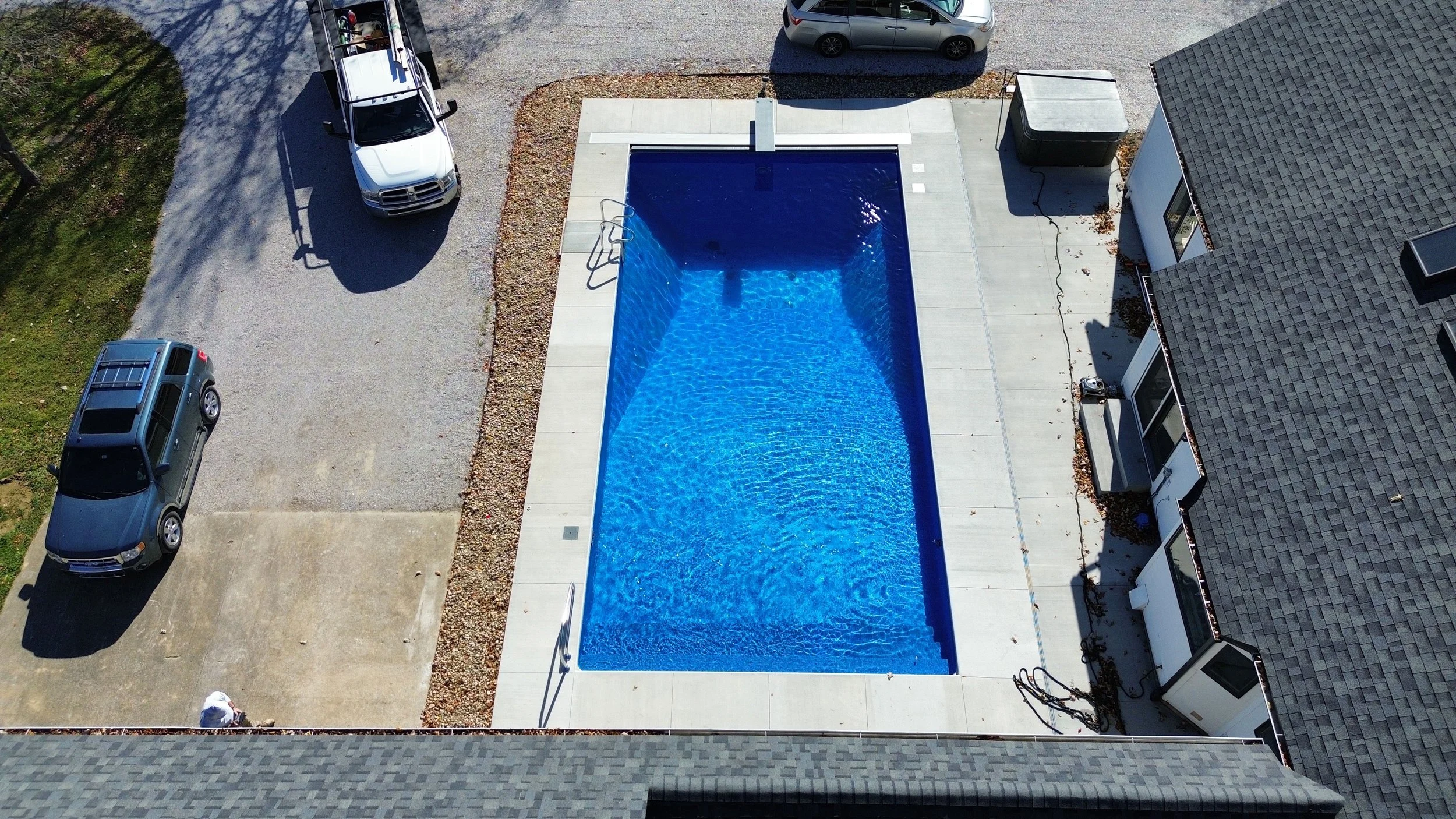An aerial view of a rectangular swimming pool with blue water, surrounded by a concrete deck, with a residential building on one side and parking spaces with cars on the other. The pool has metal ladders on one end and shadows cast by nearby structur