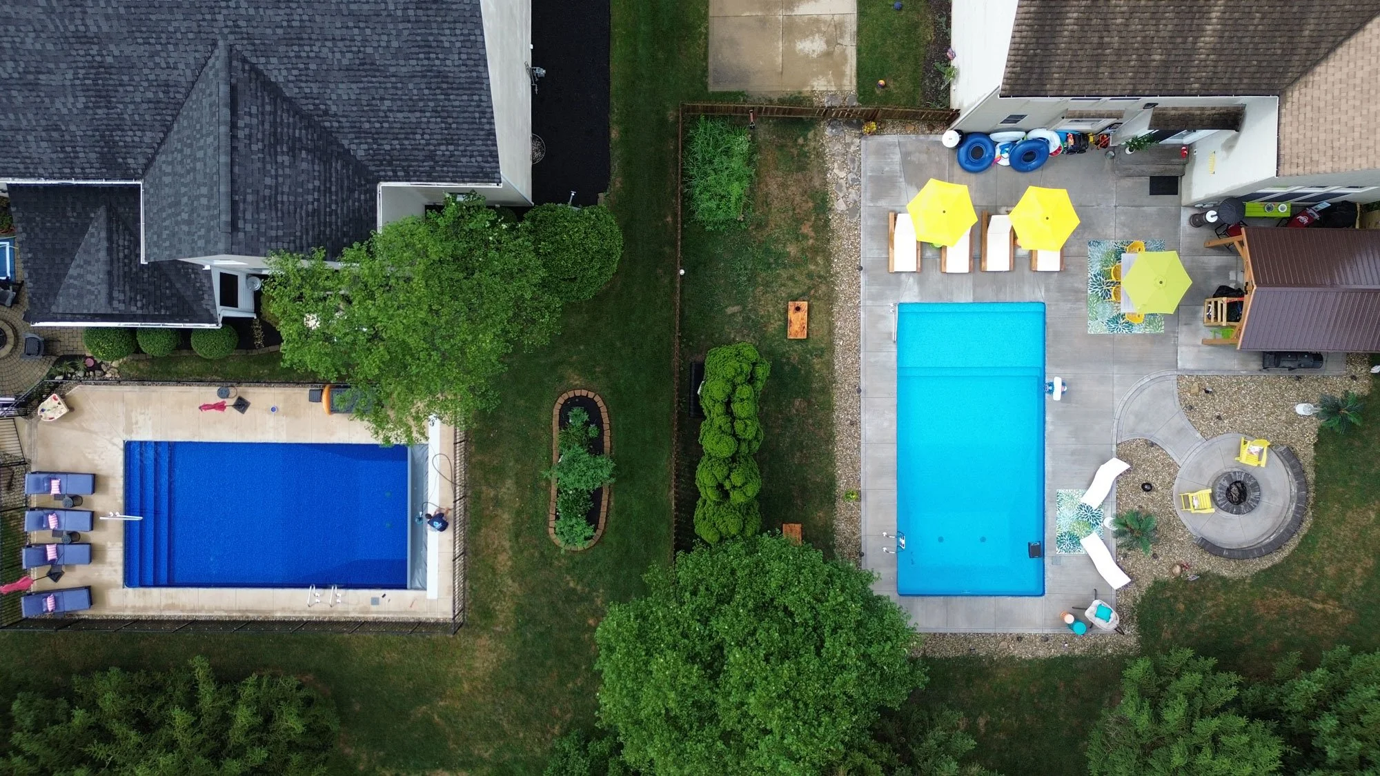 An aerial view of a backyard with two swimming pools, one with a blue cover and one uncovered. There are lounge chairs, umbrellas, and a fire pit with yellow chairs around it. The yard is divided into sections with grass, concrete, and gravel, surrou