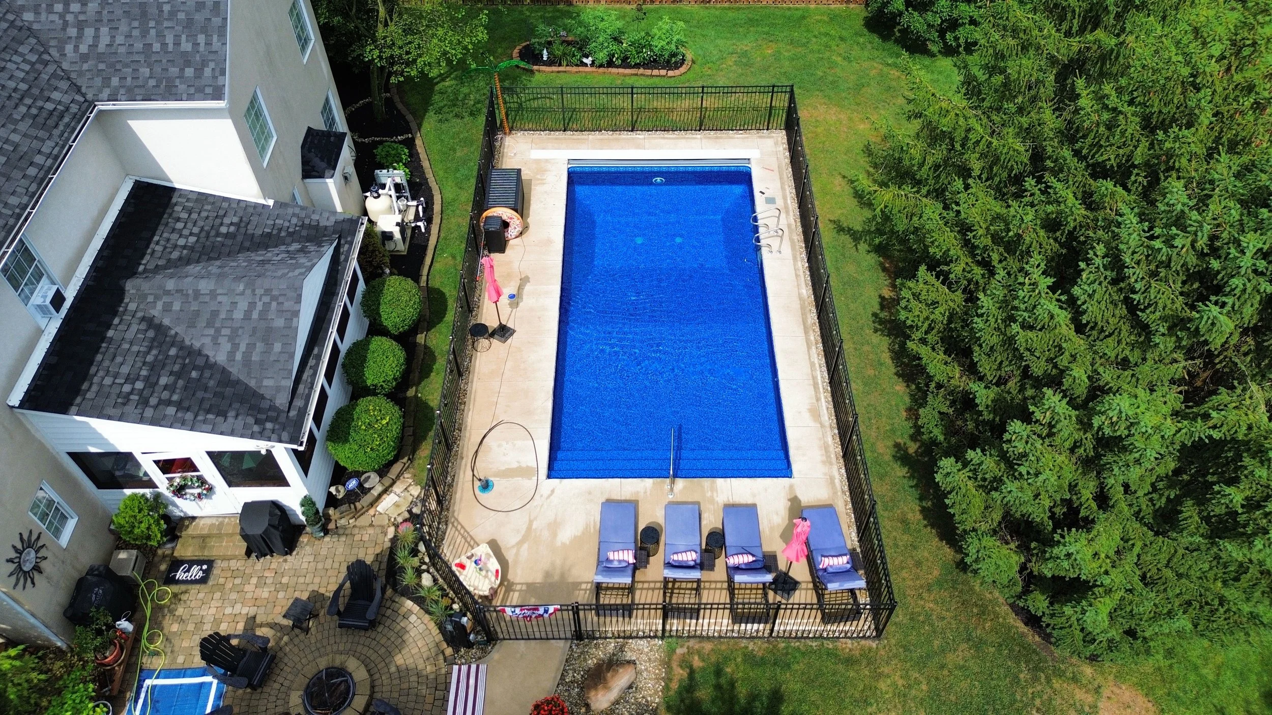 A backyard with a swimming pool, poolside chairs, and a small patio area with outdoor furniture, surrounded by greenery and trees.