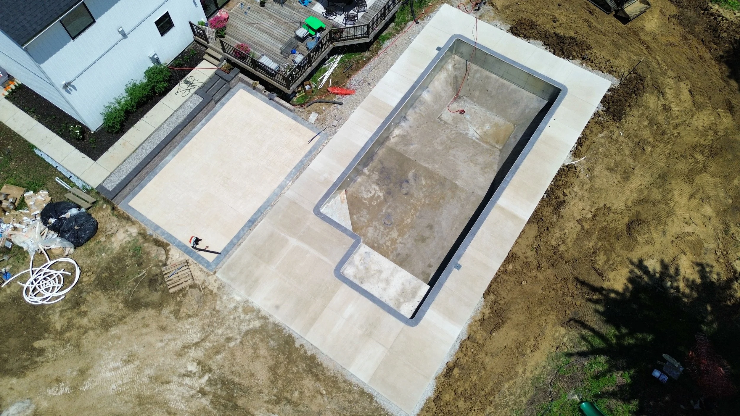 Aerial view of a backyard under construction featuring an unfilled swimming pool, a patio area, and a partially finished concrete surface, with construction materials and landscaping work in progress.