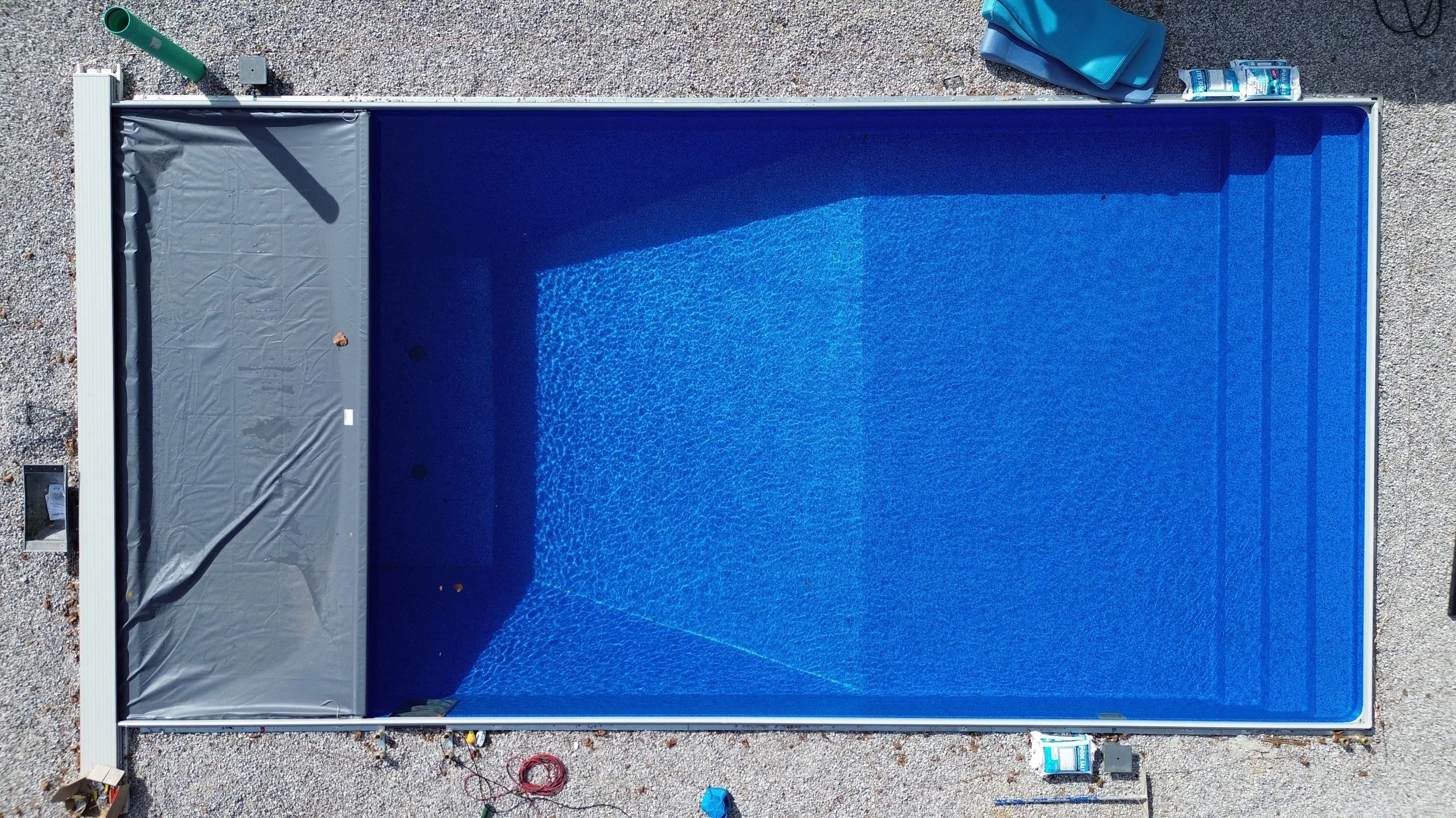 Aerial view of a rectangular swimming pool with a partially covered one end, surrounded by a gravel surface, with pool maintenance equipment and supplies nearby.