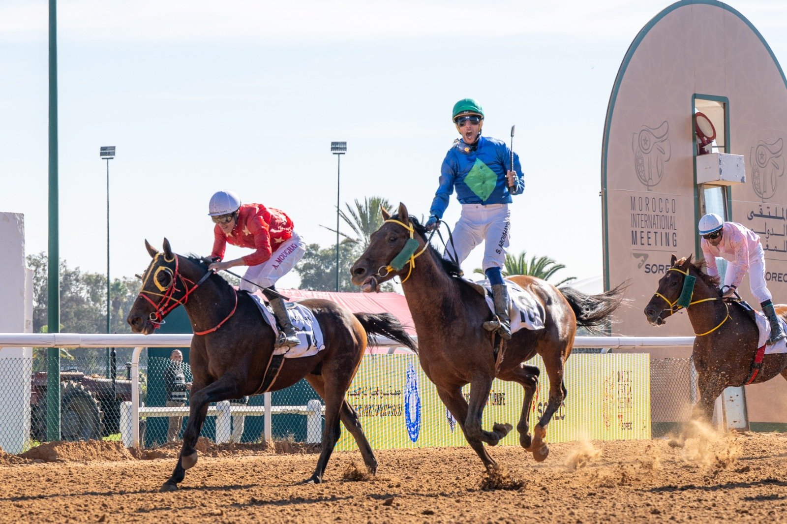 Carriles de carrera de caballos en un evento en Marruecos, con tres jinetes en el momento de la competencia, un fondo con estructuras y publicidad, y un cielo despejado.