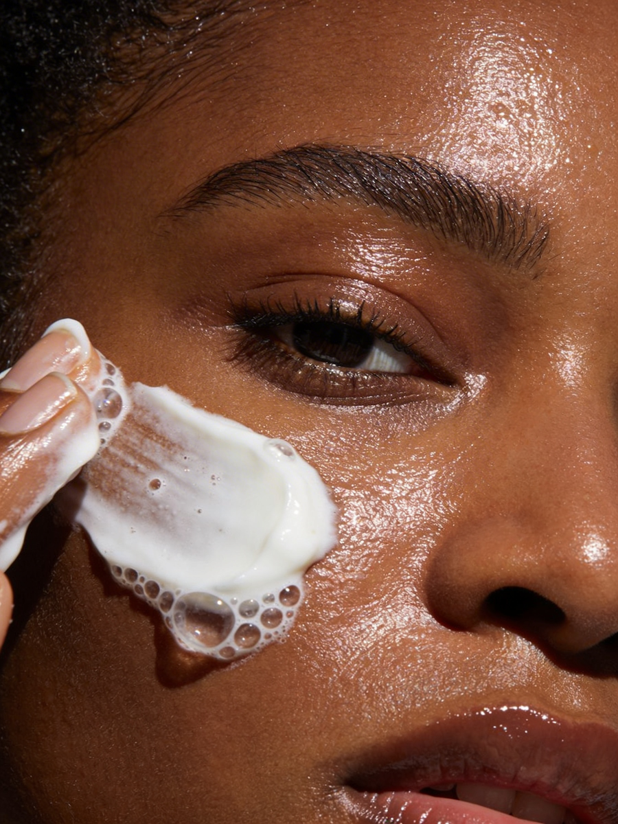 Close-up of a person with dark skin applying foamy white facial cleanser or soap to their cheek.