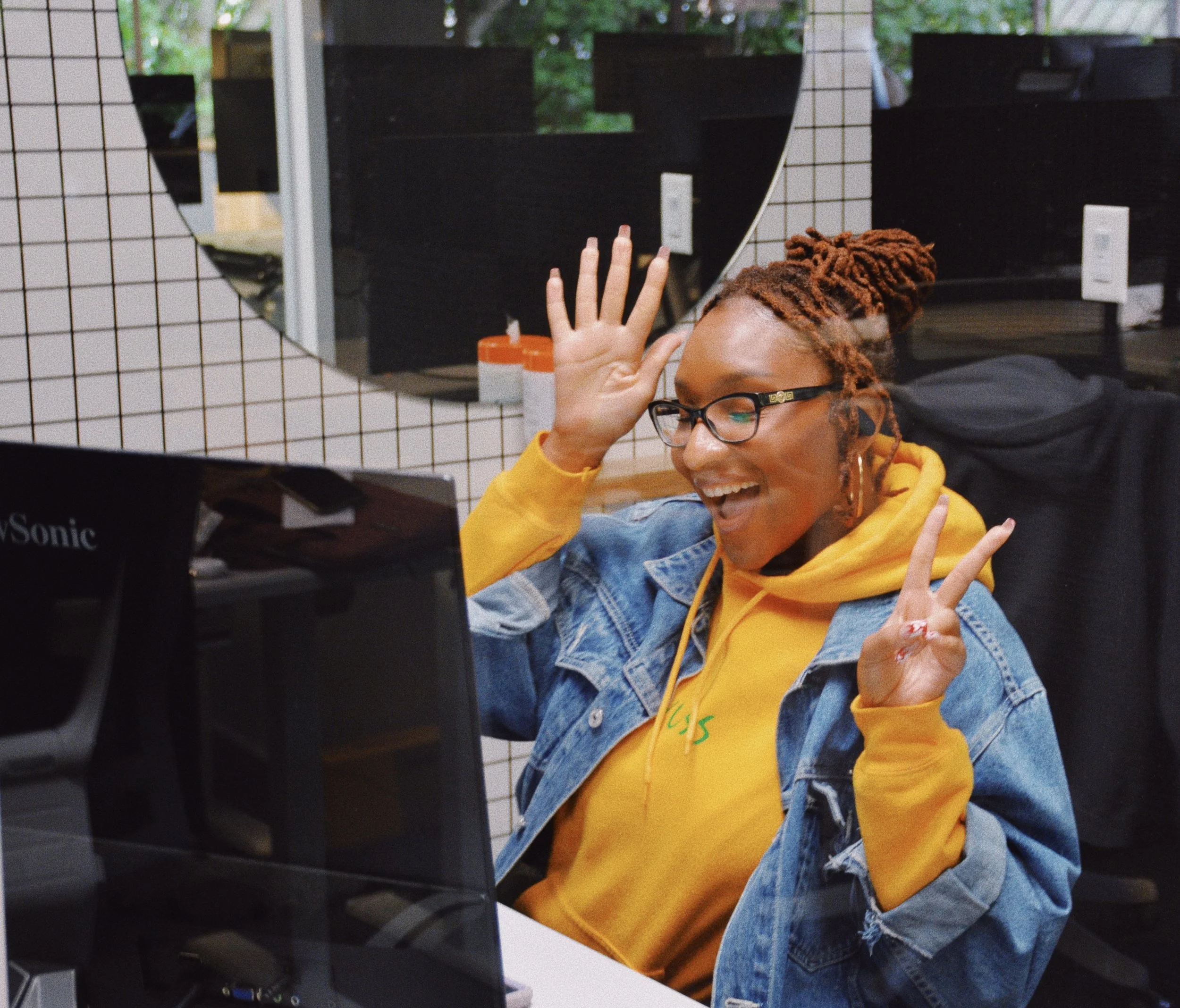 A woman with reddish-brown dreadlocks and glasses smiling and waving, sitting in front of a computer in a room with a mirror and tiled wall.