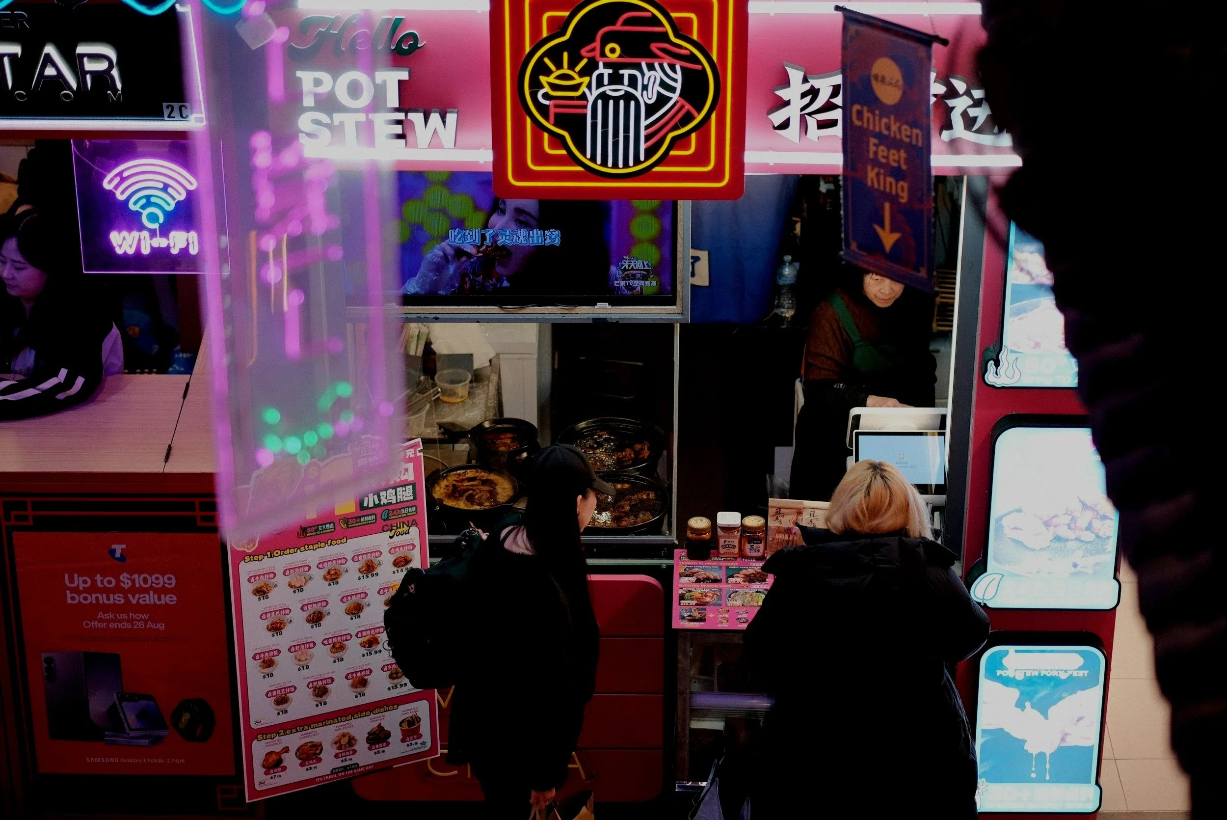A fast food restaurant stall featuring neon signs, menus, and customers ordering. Neon signs include a Wi-Fi symbol, a hot pot stew, and a stylized chicken logo. Customers are standing in front of the counter, with food and drink items visible.