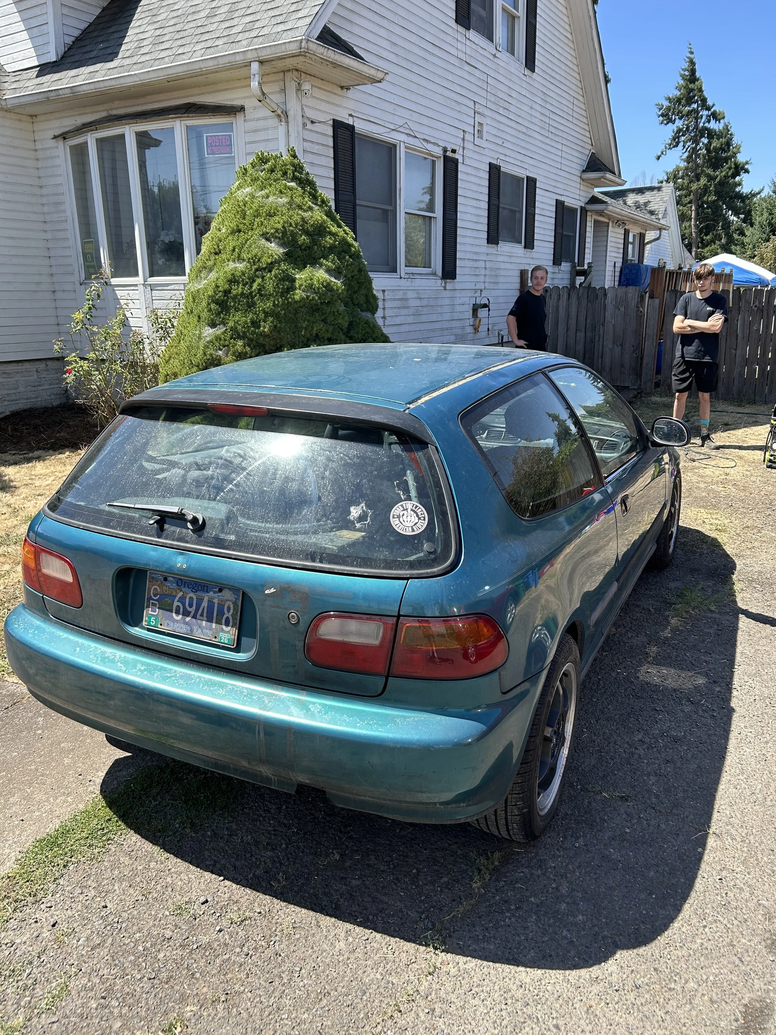 A blue hatchback car parked on a gravel driveway in front of a white house with black shutters. Two young men are standing behind the car, one with arms crossed. The house has a window with a pink sign and a large bush near the wall. The driveway is rough, with patches of grass.