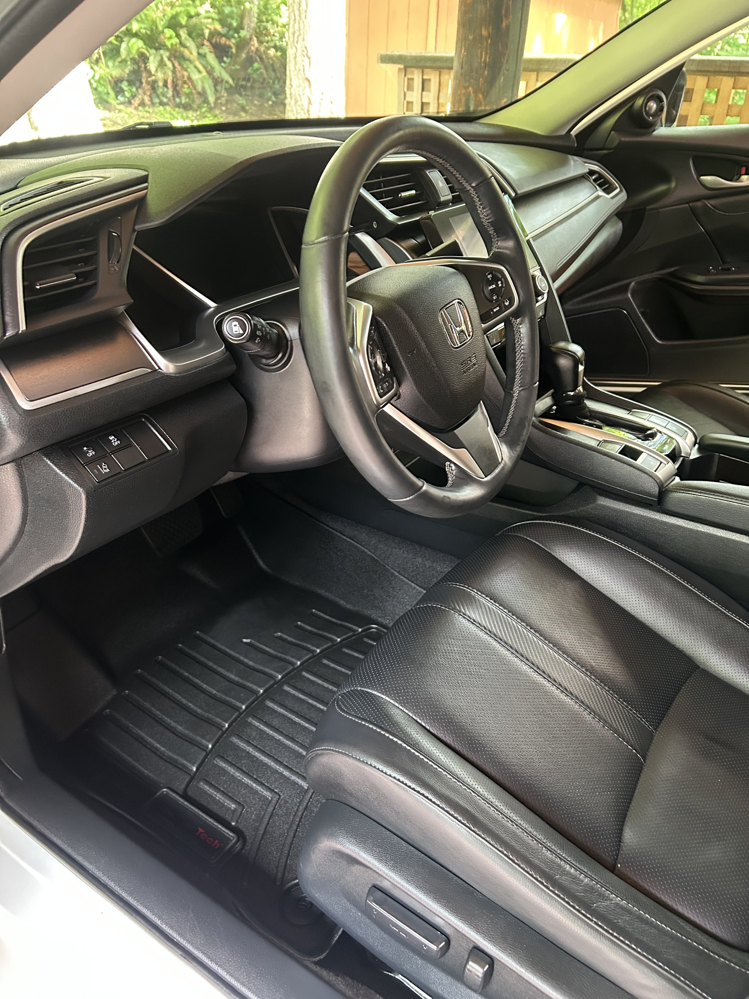 The interior of a modern car, showing the dashboard, steering wheel with Honda logo, gear shift, and black leather seats, with a view outside of a backyard with trees and a wooden fence.