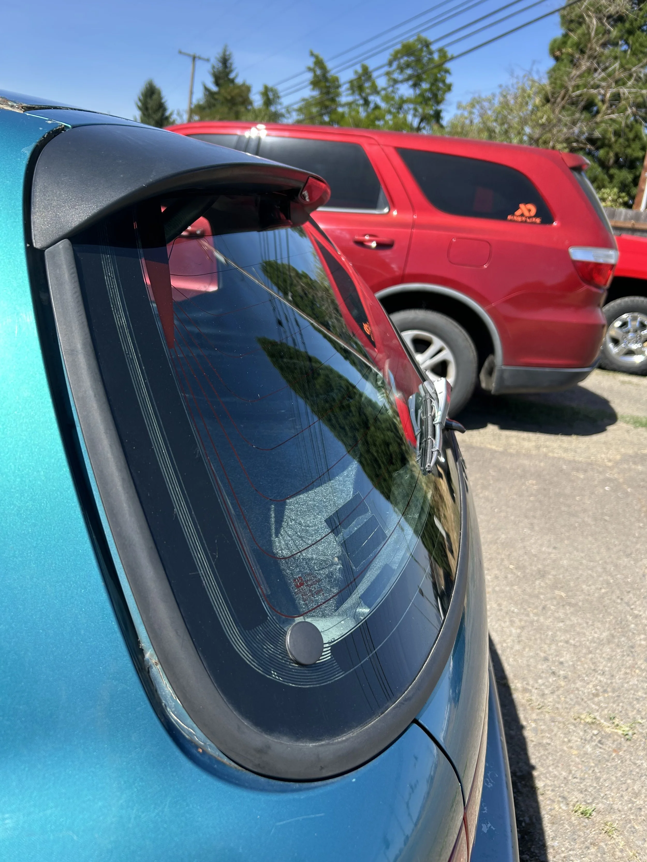Close-up of a teal-colored car's rear window with reflections of trees and power lines, and a red SUV parked next to it on a sunny day.