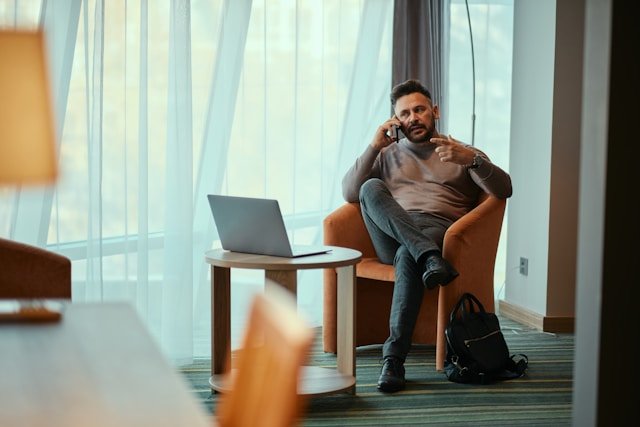 Man sitting in an orange armchair, speaking on a phone, with a laptop on a small table beside him in a hotel or office lounge area.