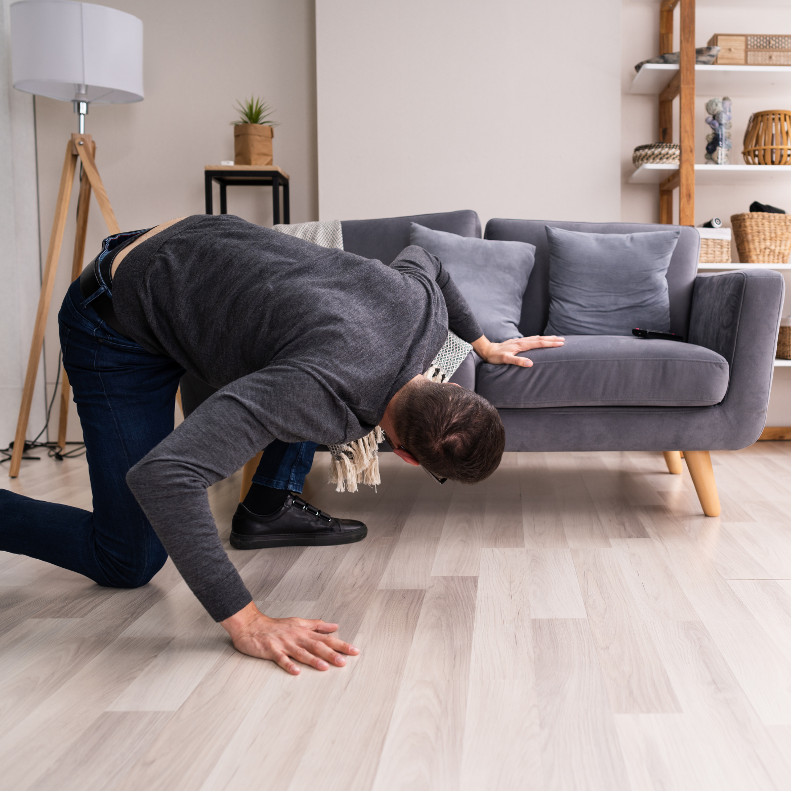 A man kneeling and leaning under a gray couch, reaching towards the floor, in a modern living room.