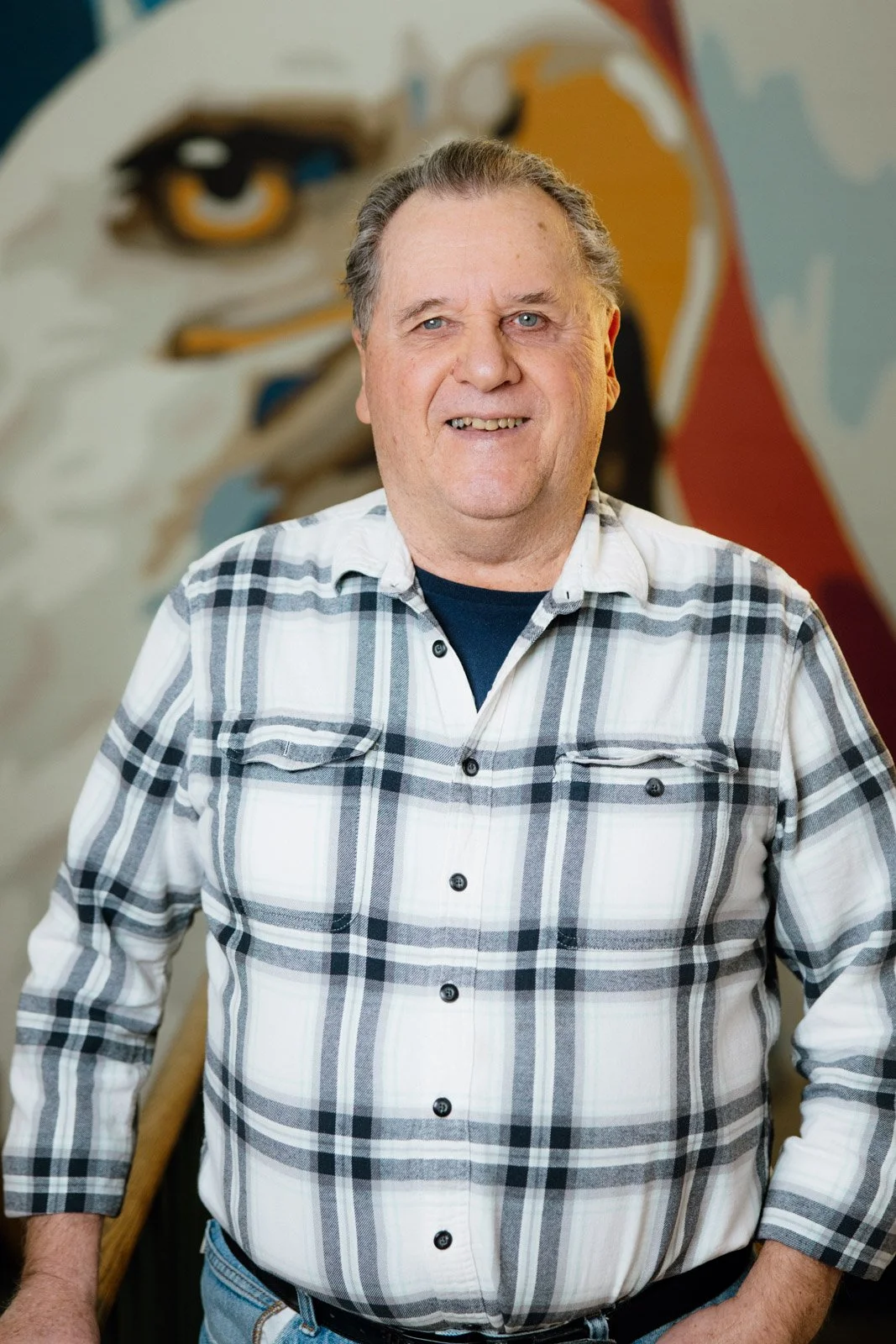 A middle-aged man with gray hair and a plaid shirt standing in front of a mural of an eagle.