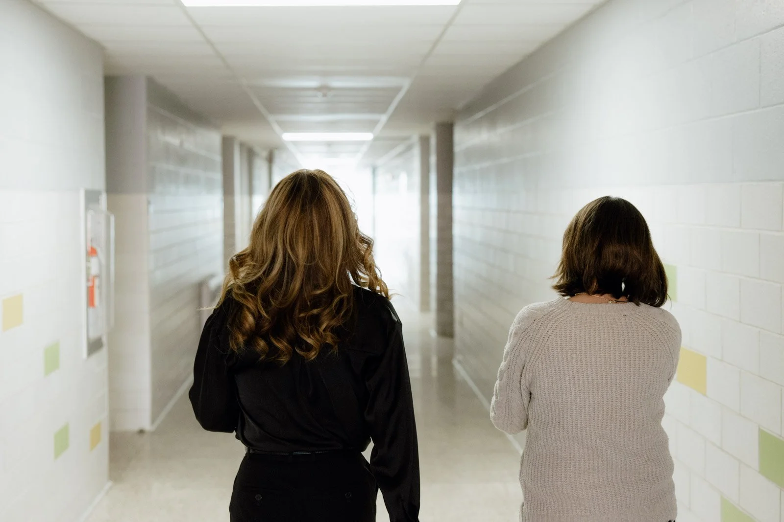Two women walking down a school hallway with white brick walls and fluorescent lighting.