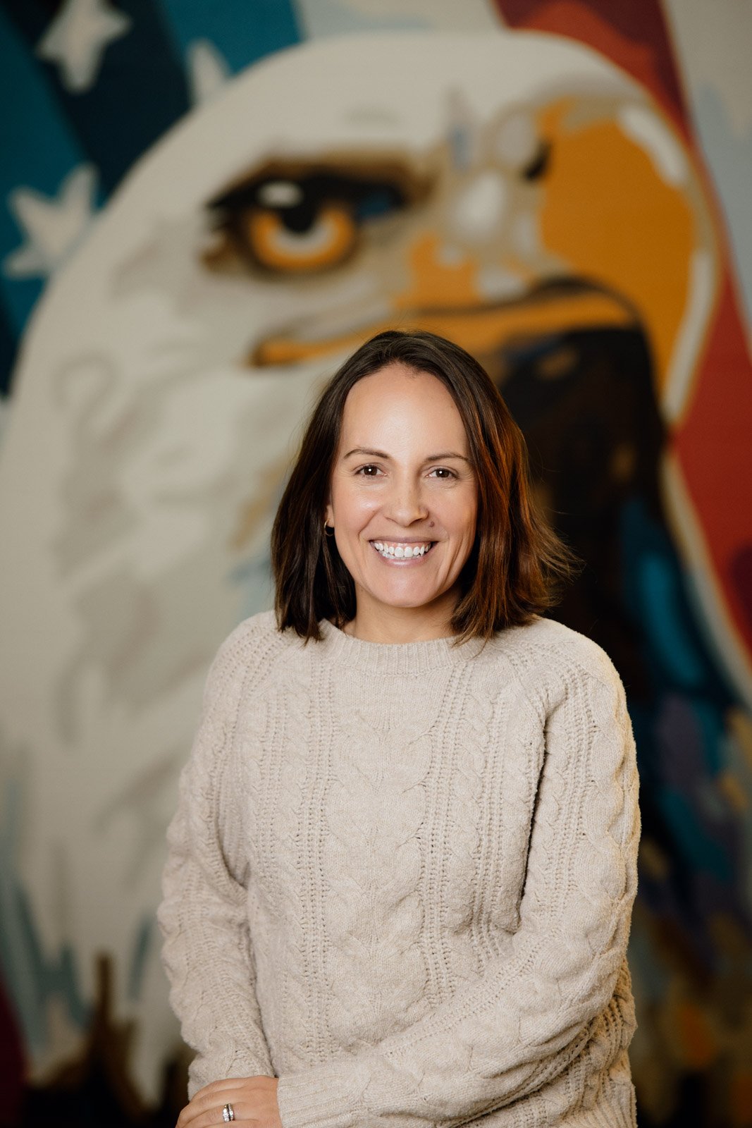 A woman with shoulder-length brown hair, wearing a beige knitted sweater, smiling while standing in front of a colorful mural of an eagle.