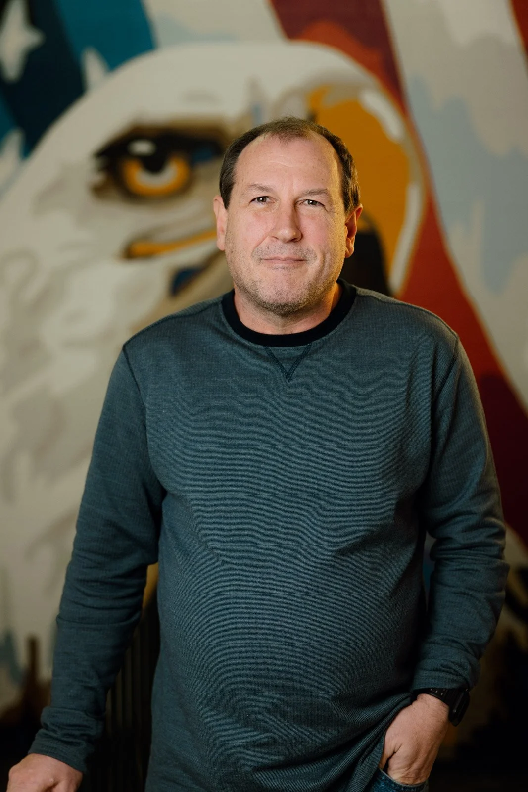 A middle-aged man with short brown hair and a beard, wearing a dark long-sleeve shirt, standing indoors in front of a colorful mural of an eagle's face.