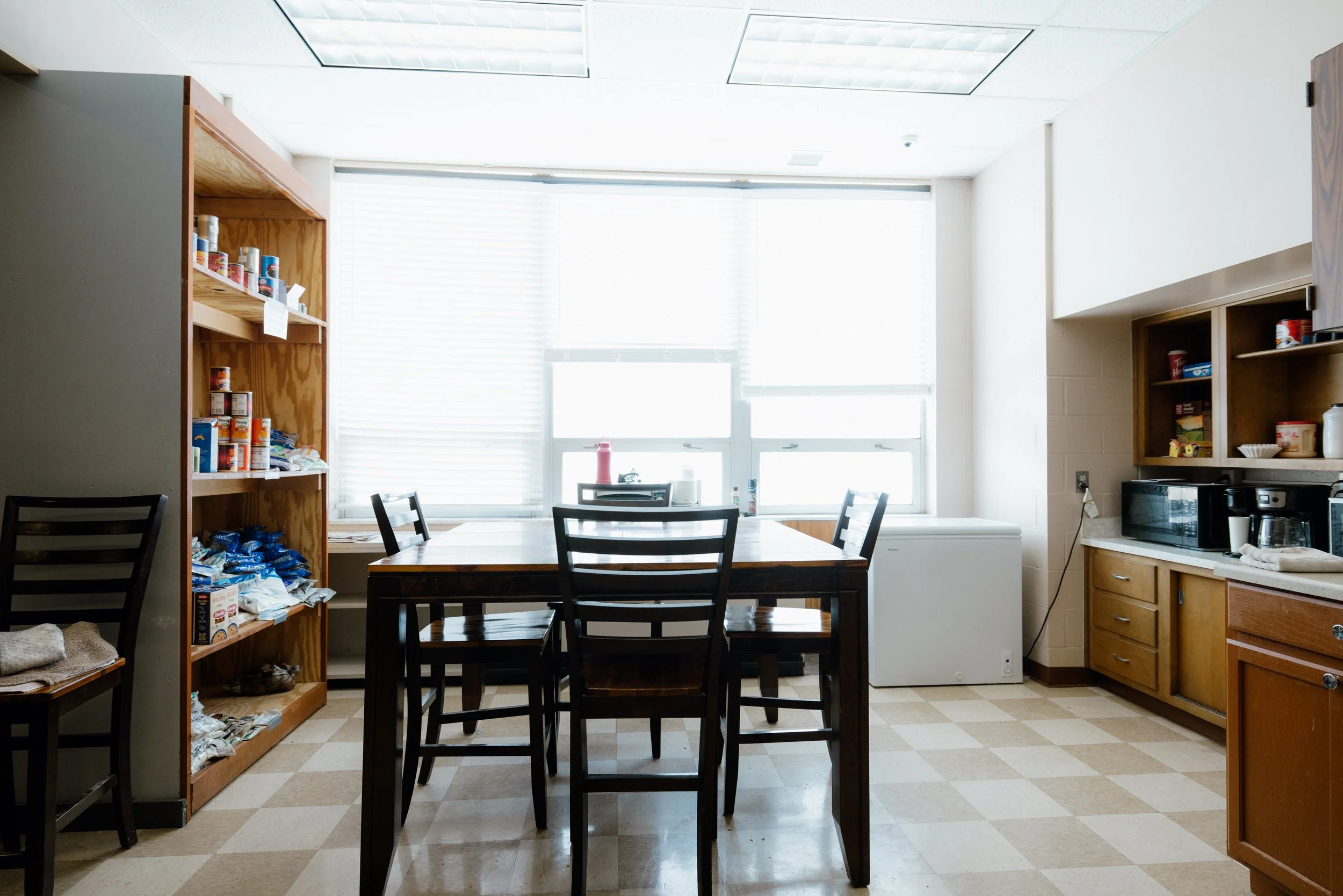 Kitchen with a wooden dining table and chairs, open shelving with canned food, a microwave, coffee maker, and various kitchen items, bright window with blinds.