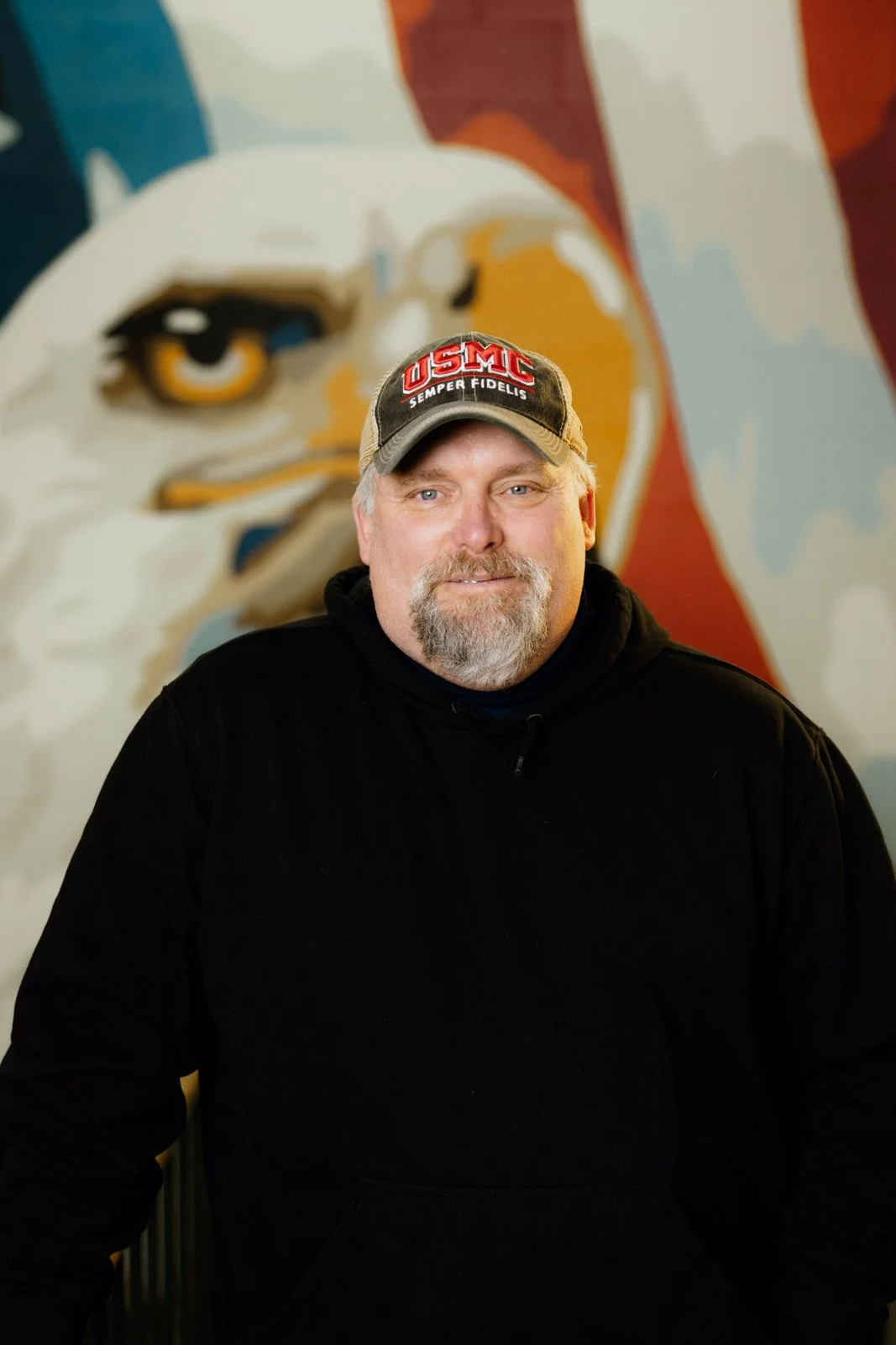 A man with a beard wearing a black hoodie and a baseball cap with 'USMC Semper Fidelis' printed on it, standing in front of a mural featuring a large eagle face.