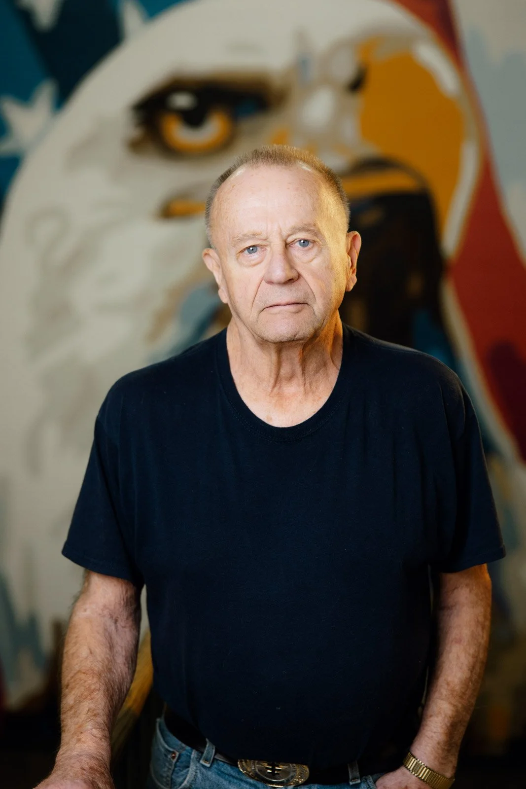 An elderly man with a serious expression, wearing a black T-shirt and a gold bracelet, standing in front of a large colorful mural of a woman's face with bold lines and vibrant colors.