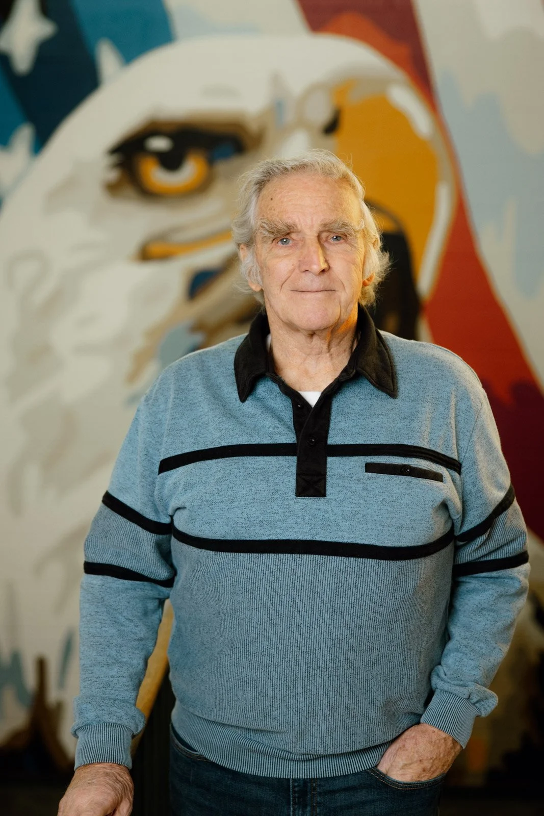 An older man with gray hair and blue eyes standing indoors in front of a mural of a stylized eagle. He is wearing a light blue sweater with black stripes and a black collar shirt underneath. His right hand is in his pocket and he has a neutral expression.