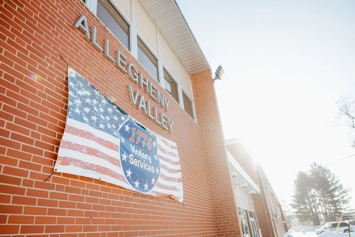 Exterior of a brick building with signs that read "Allegheny Valley" above and a patriotic banner with the American flag and emblem that says "1776 Notary Services" hanging on the wall. The sun shines brightly, creating lens flare, and trees and parked cars are visible in the background.