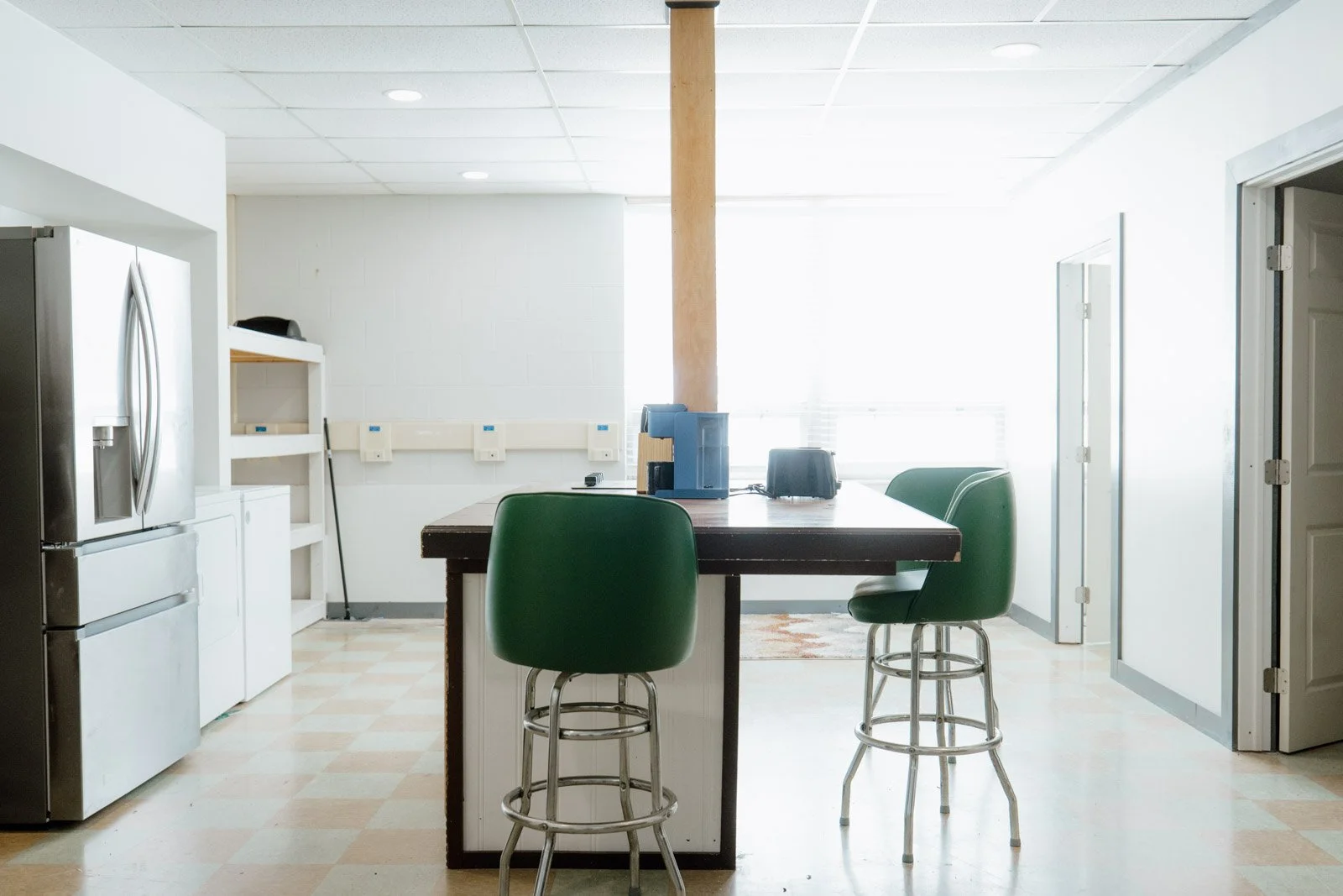 Empty kitchen with a black island counter, green chairs, stainless steel appliances, and minimal decor, illuminated by natural light from a window.