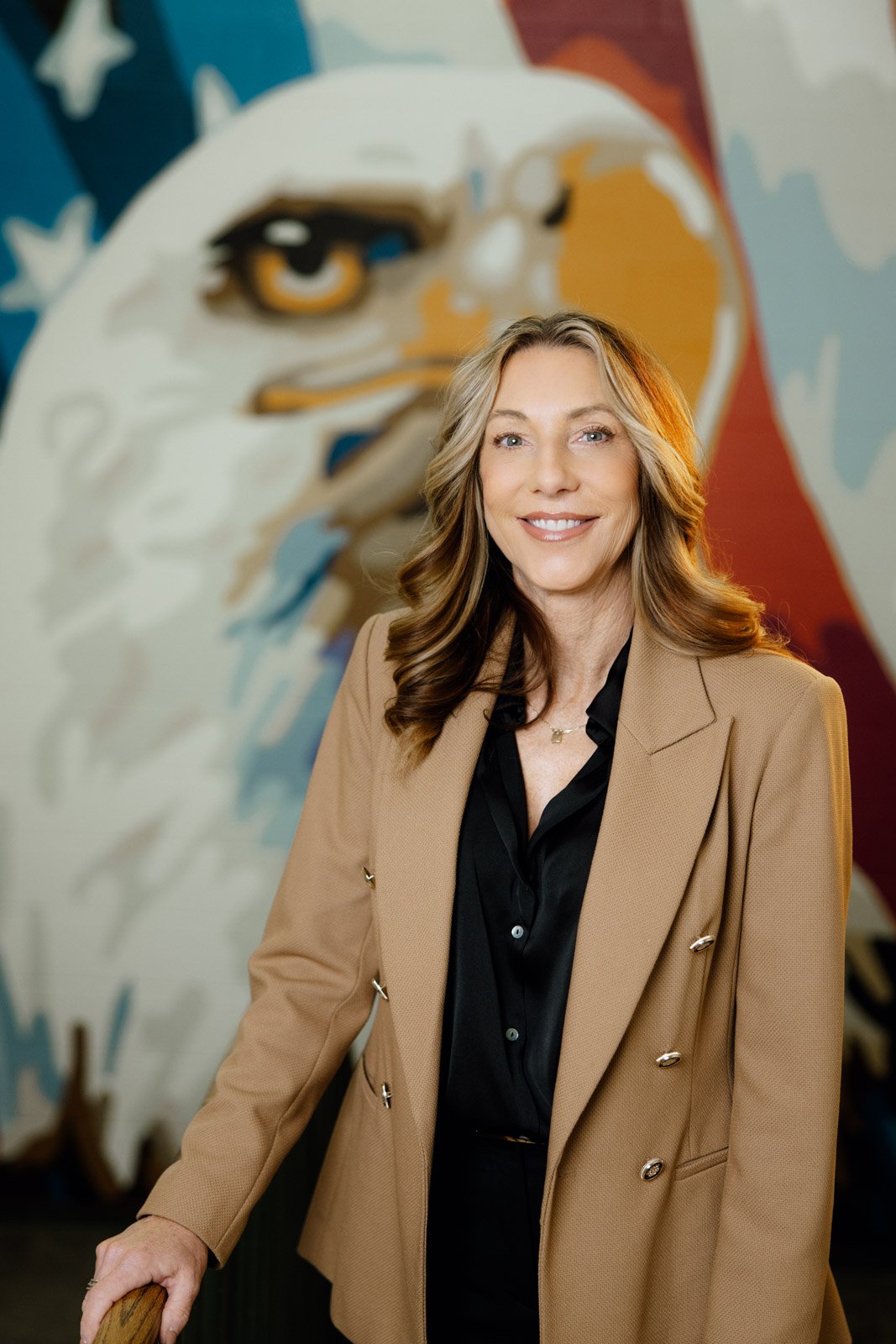 A woman with wavy light brown hair, wearing a tan blazer over a black blouse, standing in front of a large mural of a bald eagle.