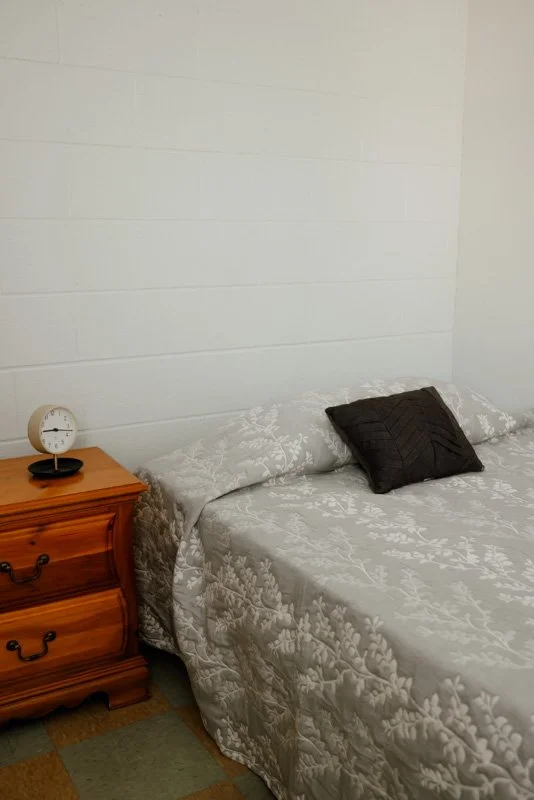 A bedroom with a bed with a floral quilt and a black pillow, a bedside wooden nightstand, and a small clock on the nightstand.