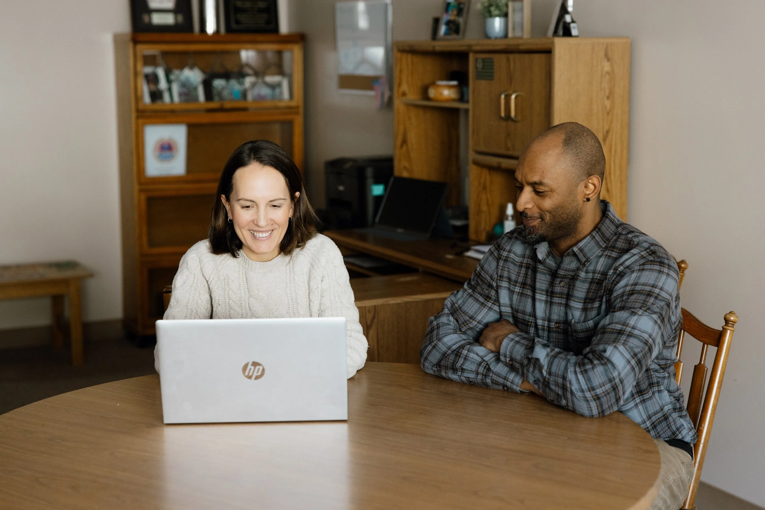A woman and a man sitting at a wooden table looking at a laptop, smiling, in a home office with wooden furniture and bookshelves in the background.