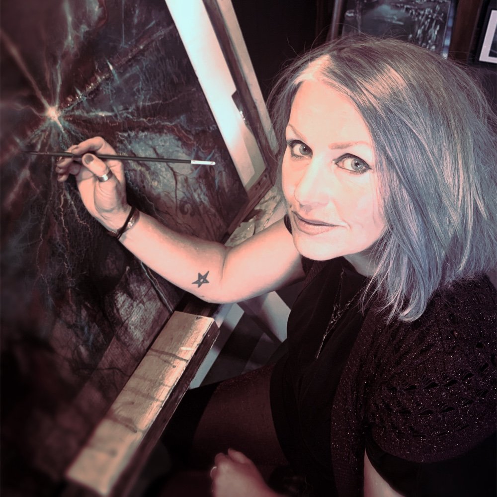 A woman with shoulder-length gray hair painting a landscape on a canvas. She is sitting at a table, holding a paintbrush in her right hand, and looking up at the camera. She has a star tattoo on her left wrist and is wearing a black top.