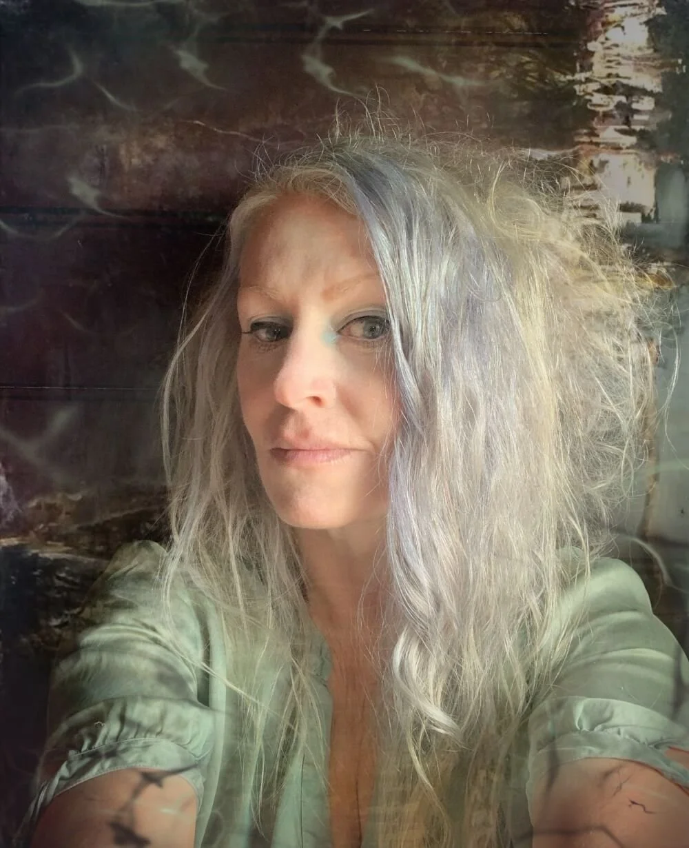 A woman with long, frizzy, gray hair taking a selfie in front of a dark, marbled background.