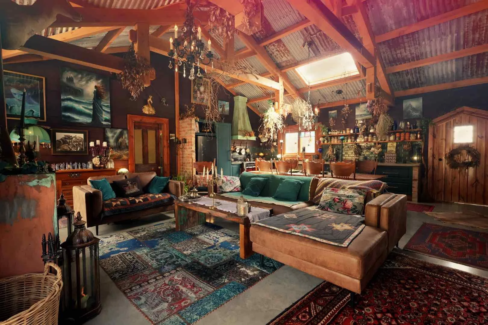 A cozy, rustic living room with vintage decor, colorful patchwork rugs, plush sofas with pillows, wooden furniture, and an open kitchen area in the background. The room has warm lighting and decorative chandeliers hanging from the ceiling.
