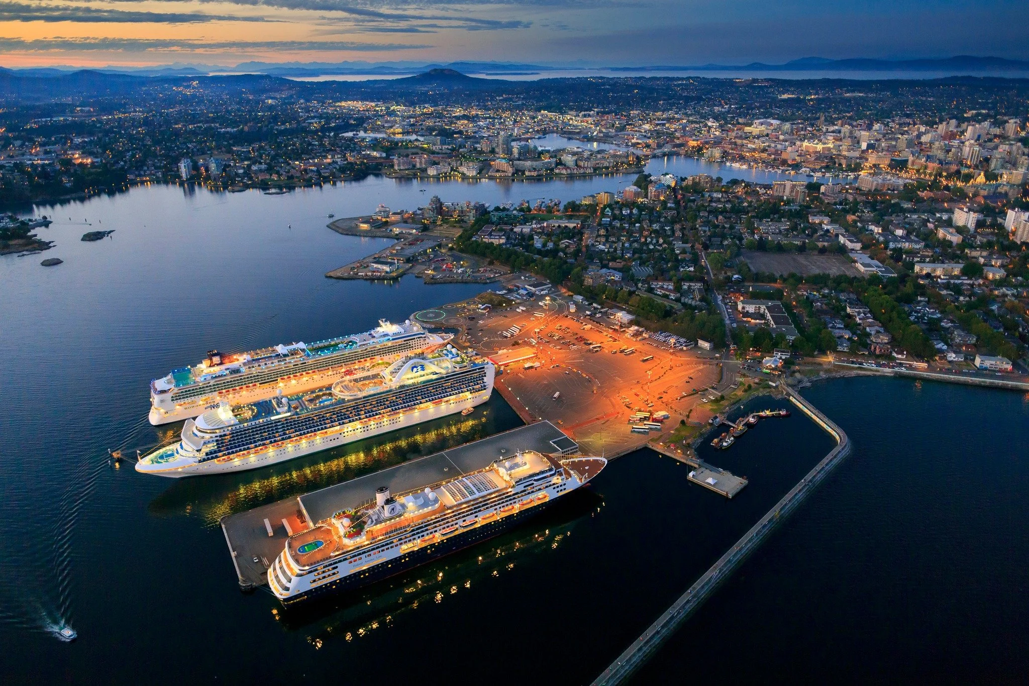 Aerial view of Victoria, BC harbor at dusk with three illuminated cruise ships docked, surrounding parking lots, and a cityscape in the background showing buildings and a river.