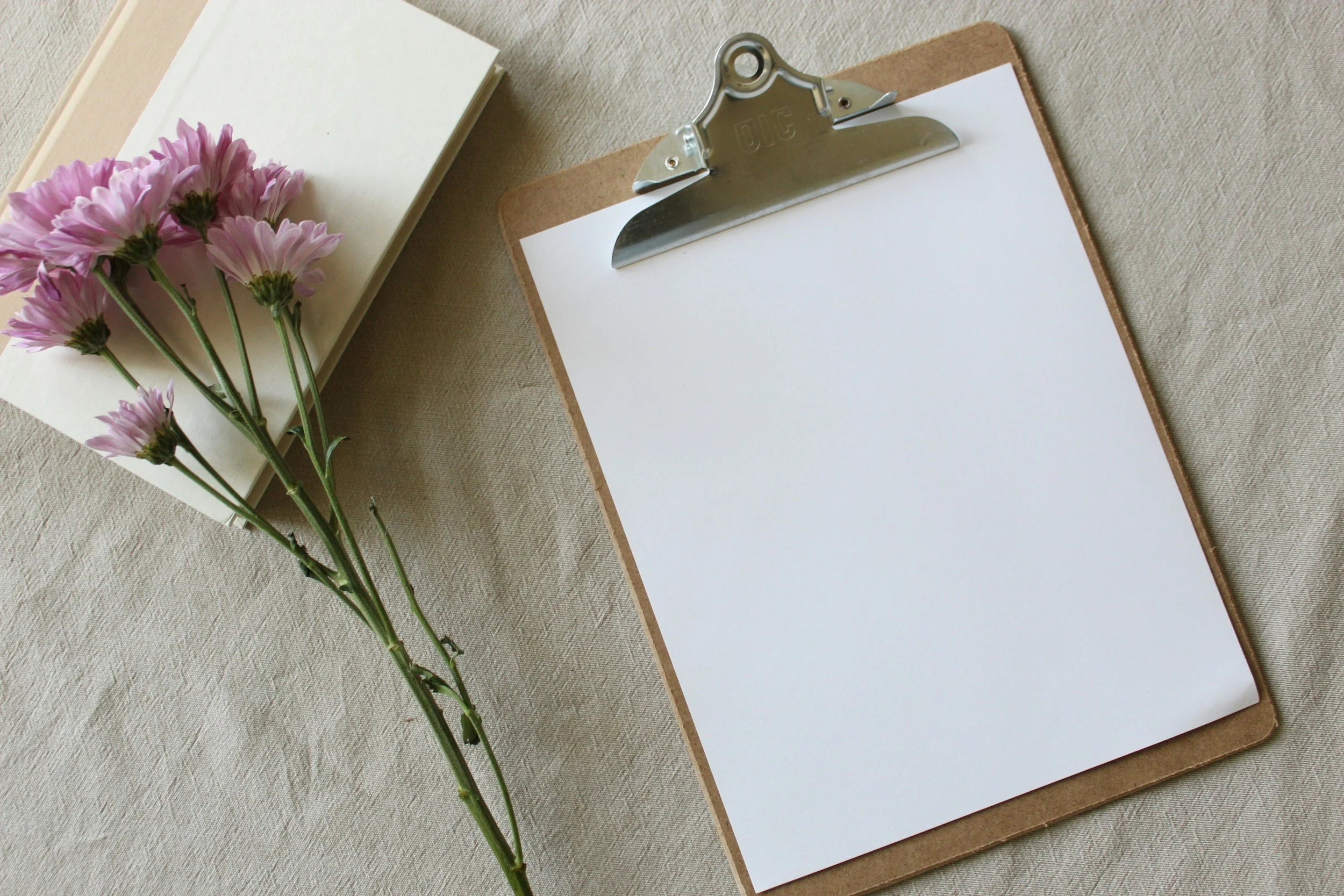 A clipboard with a blank sheet of paper, a bunch of pink flowers, and a beige notebook on a light fabric surface.