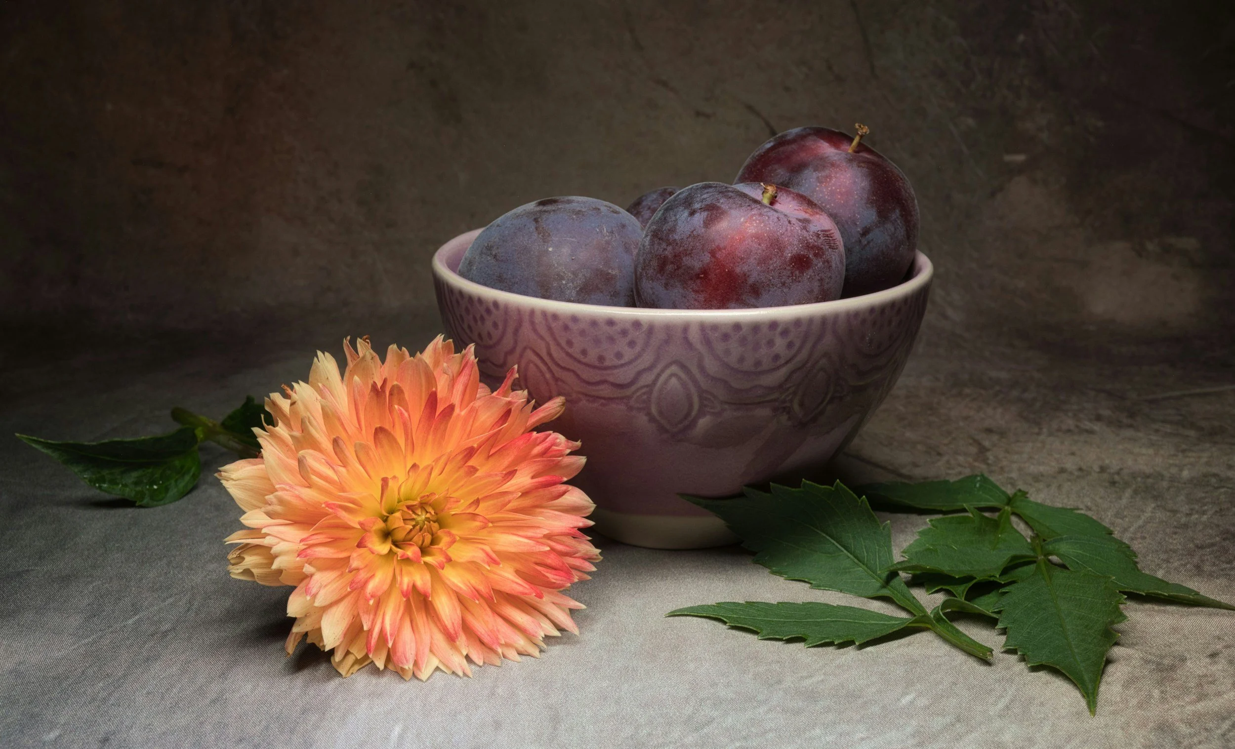 A bowl of purple plums with a peach-colored dahlia flower and green leaves on a textured surface.