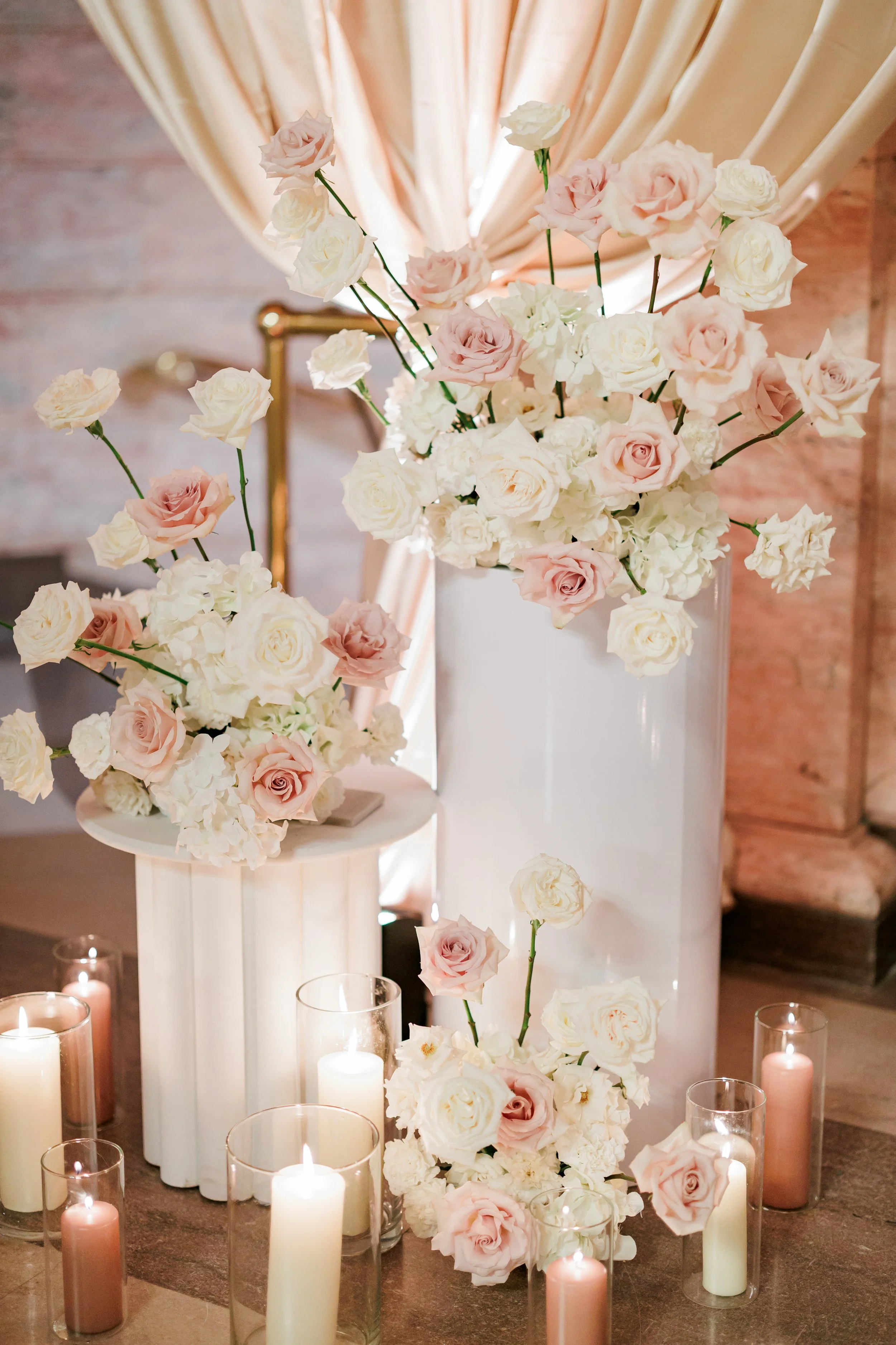 Large bouquet of white and light pink roses and hydrangeas in a white vase, surrounded by smaller arrangements and candles in glass holders, set against a background of beige curtains and a brick wall.