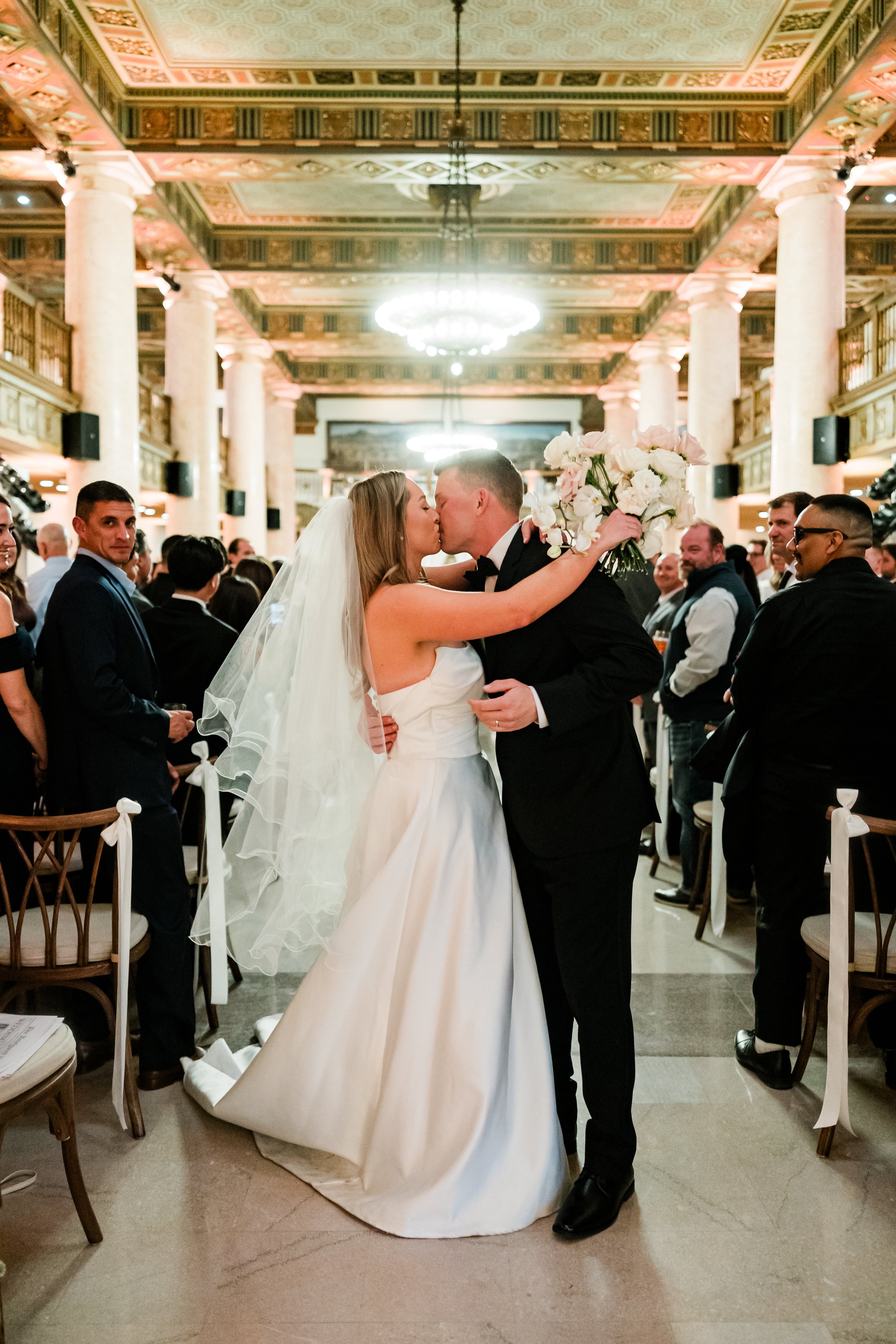 A bride and groom kiss during their wedding ceremony in a grand, ornate hall, surrounded by guests seated on chairs decorated with white ribbons, with tall columns, chandeliers, and decorative ceiling in the background.