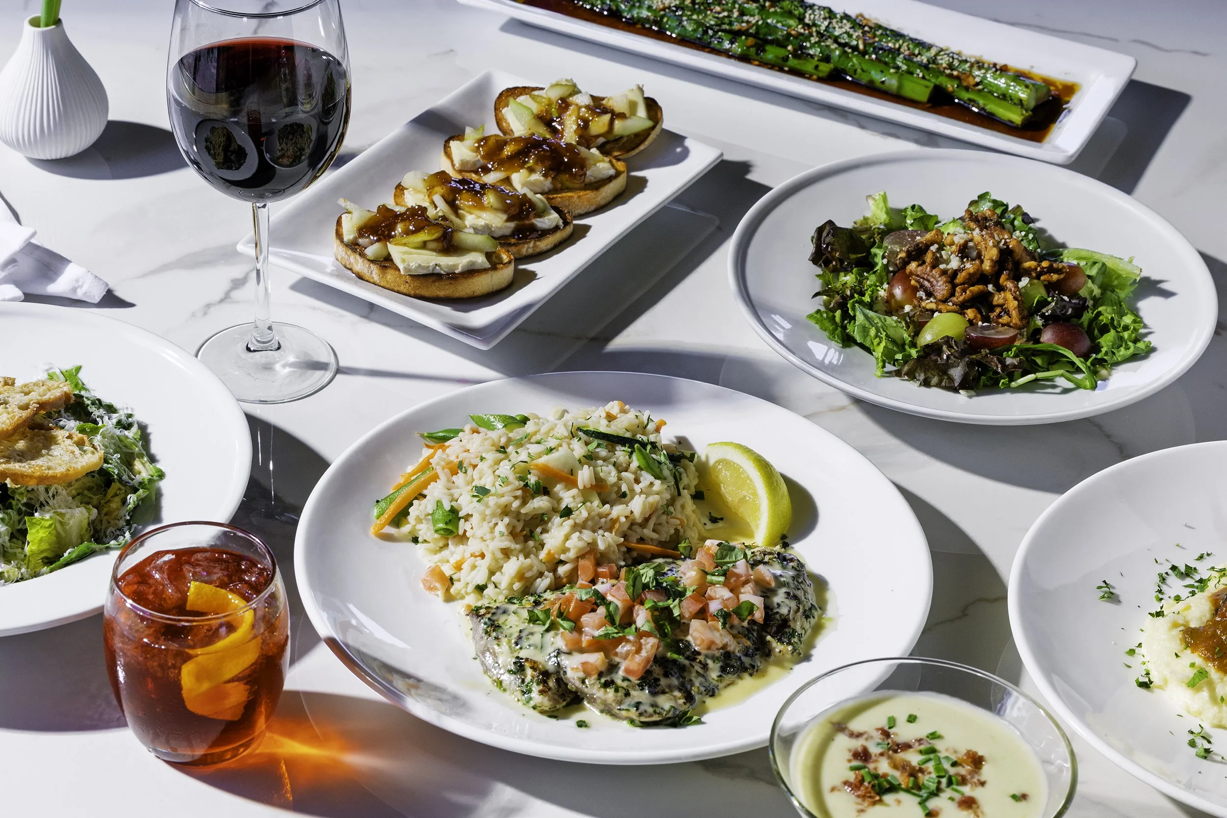 A variety of dishes on a white table, including a glass of red wine, a salad, rice with vegetables and fish, garlic bread with cheese and sauce, and a bowl of creamy dip.
